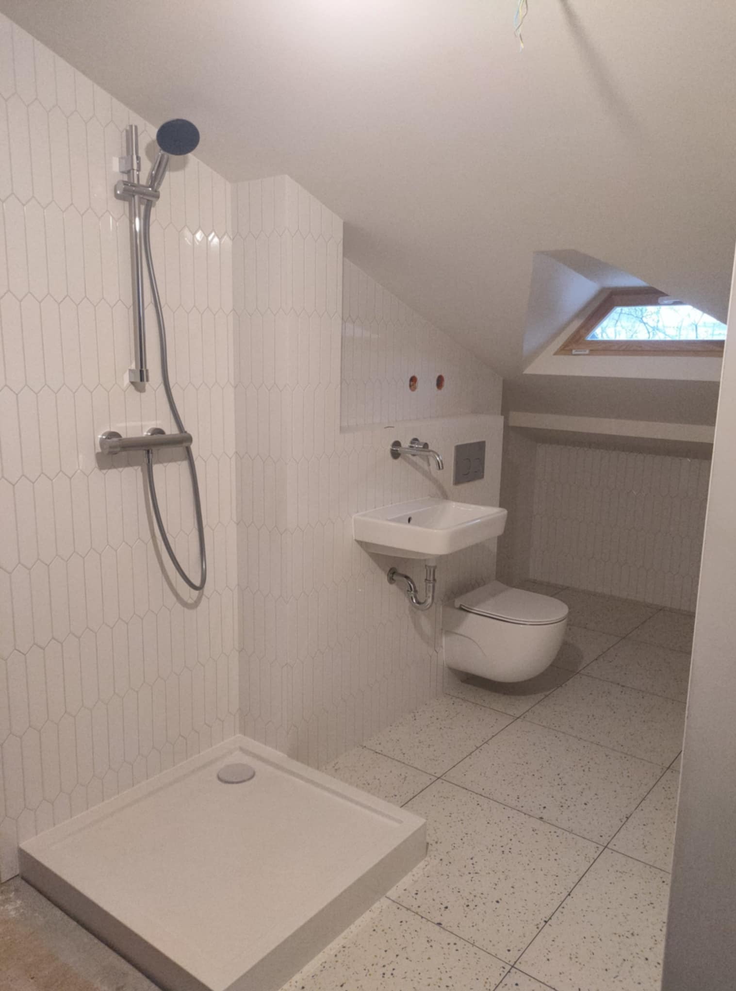 Nowoczesna łazienka na poddaszu z białą armaturą i geometrycznymi płytkami. Widoczny prysznic, umywalka i toaleta. Okno dachowe doświetla wnętrze.
