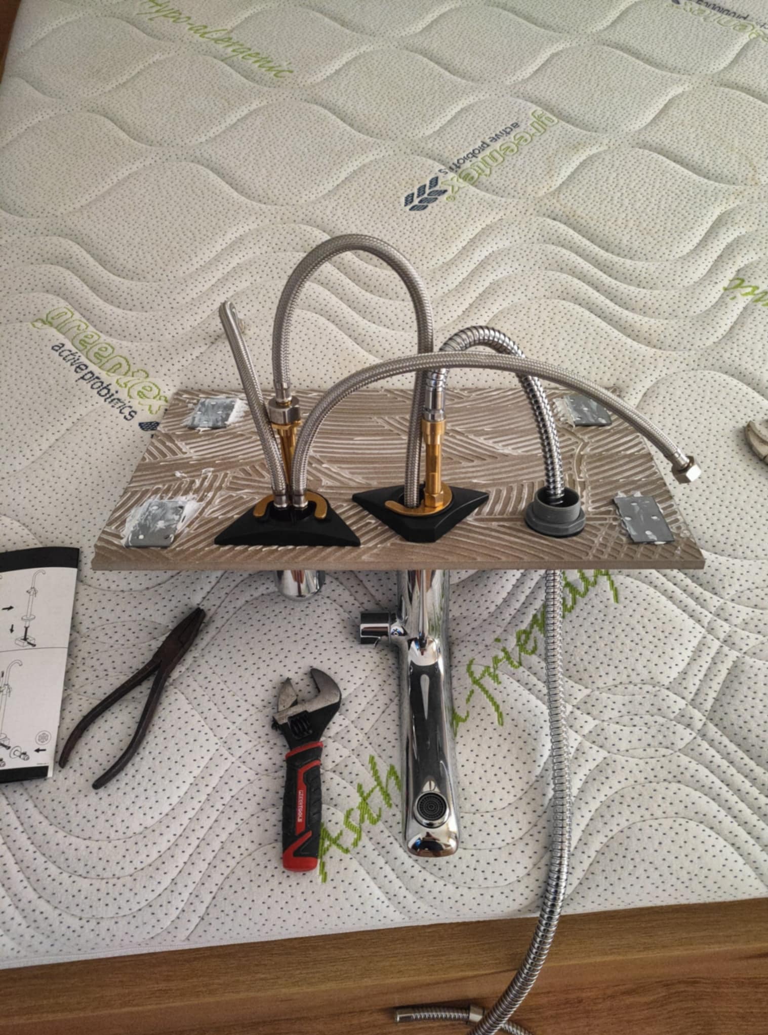 Bateria umywalkowa z wężykami i narzędziami na beżowej płytce imitującej drewno, leżącej na materacu. Widoczne szczypce i klucz nastawny.