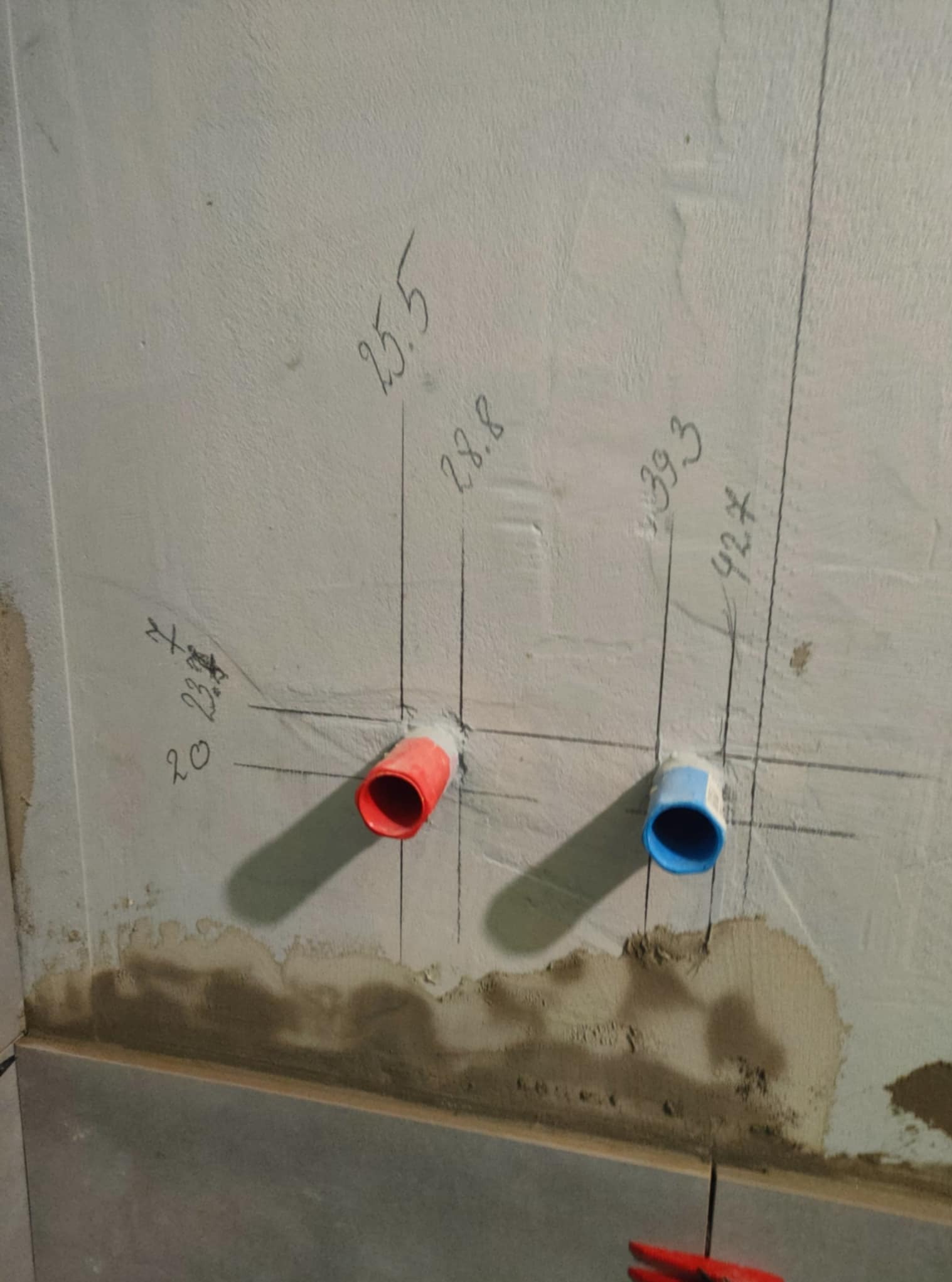 Ściana z instalacją wodną: czerwona i niebieska rurka, oznaczenia wymiarowe. Widoczne ślady po szpachlowaniu. Fragment płytek na dole kadru.