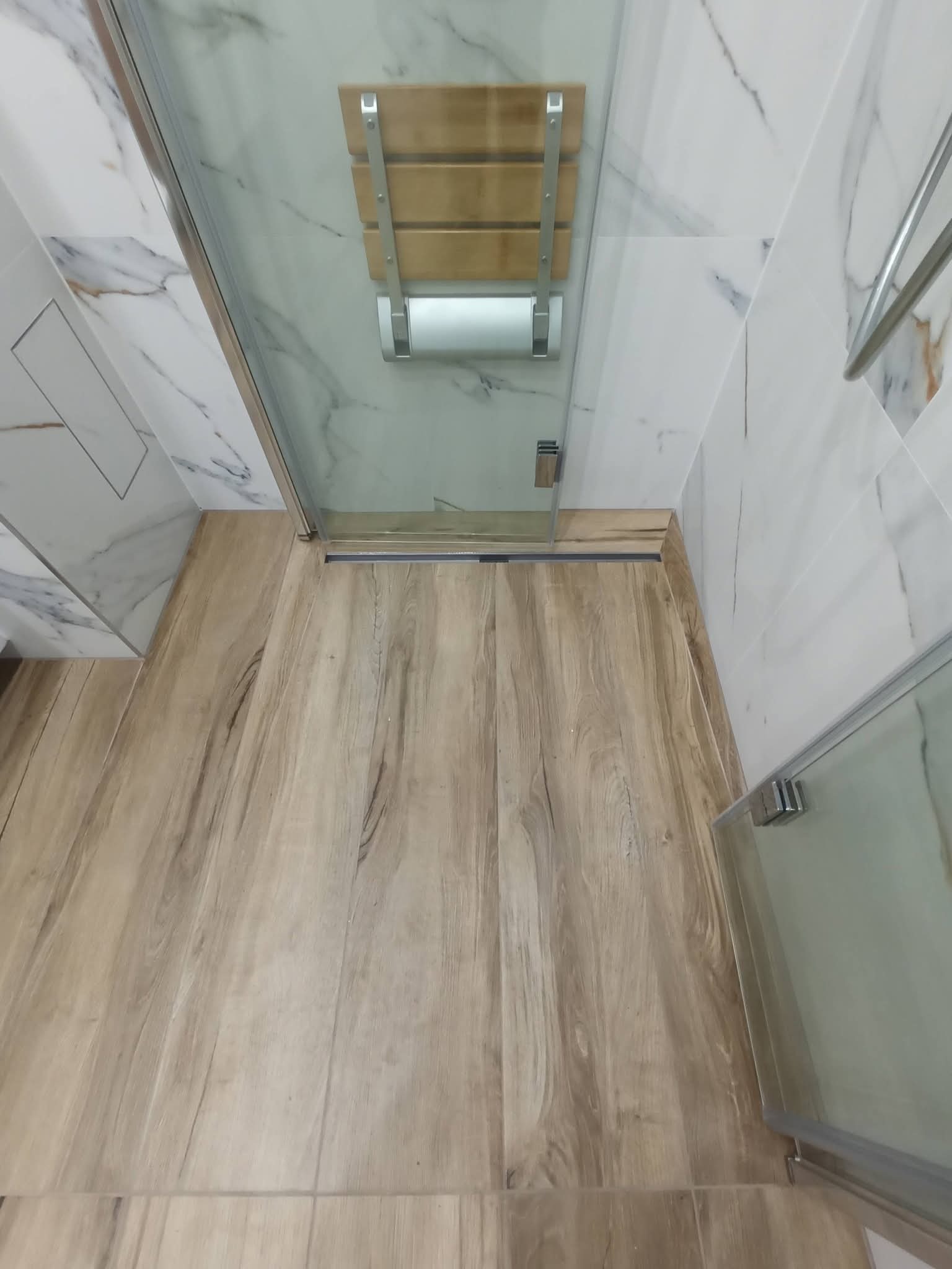 Nowoczesna kabina prysznicowa z drewnianą ławeczką i odpływem liniowym, wykończona płytkami imitującymi drewno i marmur. Widok z góry.