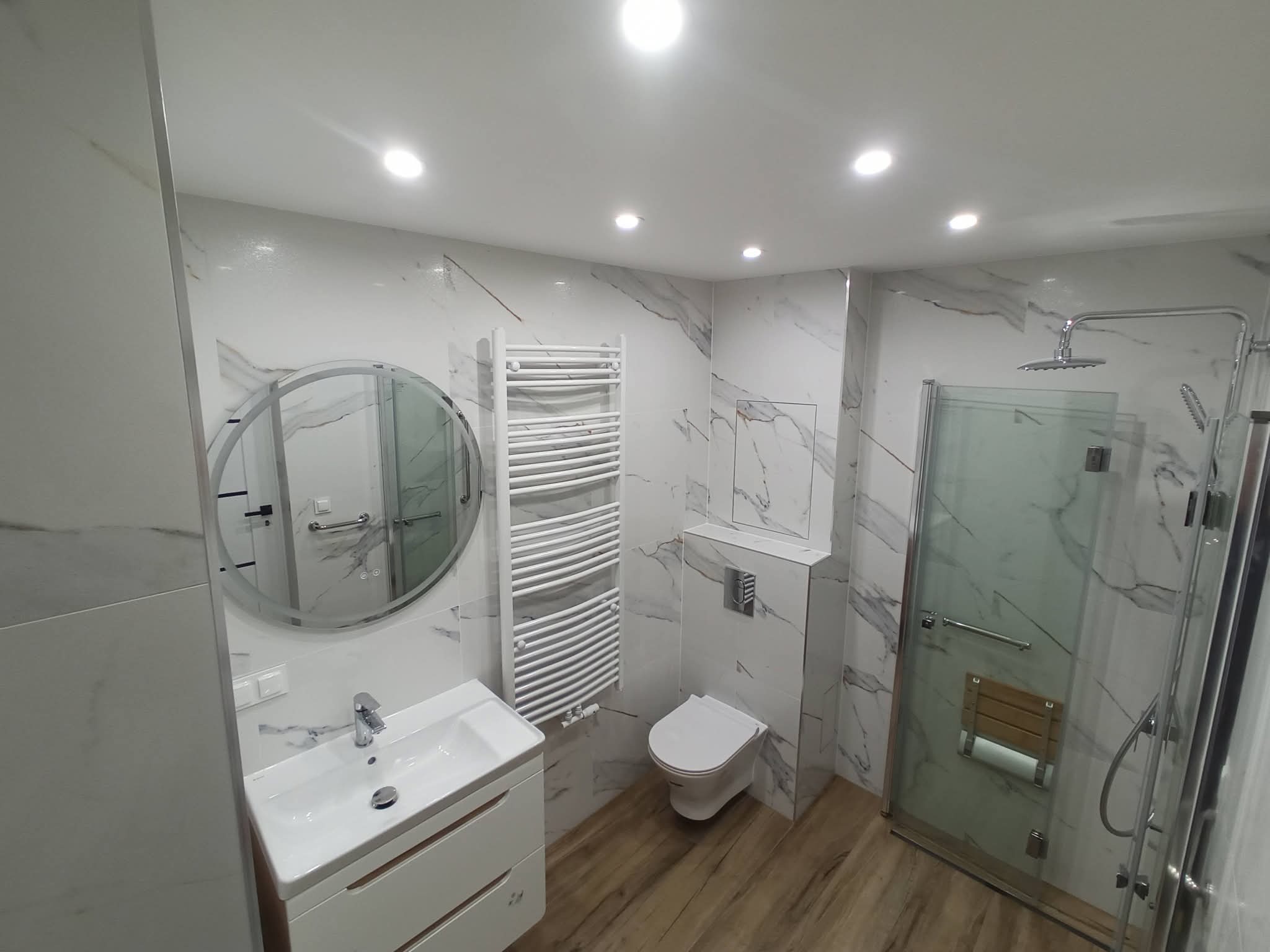 Nowoczesna łazienka z marmurowymi płytkami, kabiną prysznicową ze szklanymi drzwiami, okrągłym lustrem i drewnianą podłogą. Minimalistyczny design.