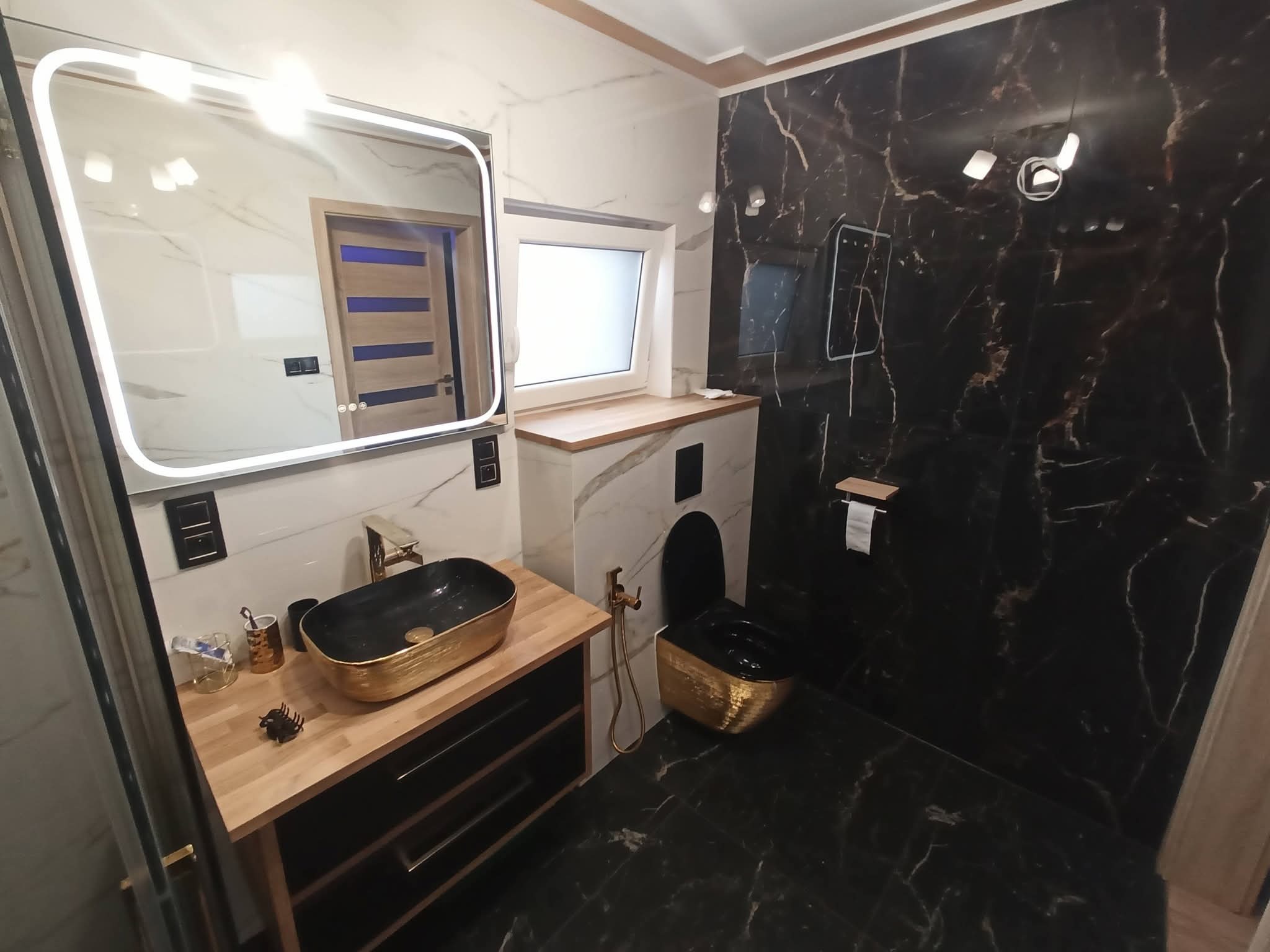 Elegancka łazienka w czerni i złocie: umywalka nablatowa, toaleta podwieszana, lustro z podświetleniem LED, marmurowe płytki. Nowoczesny design z akcentami glamour.