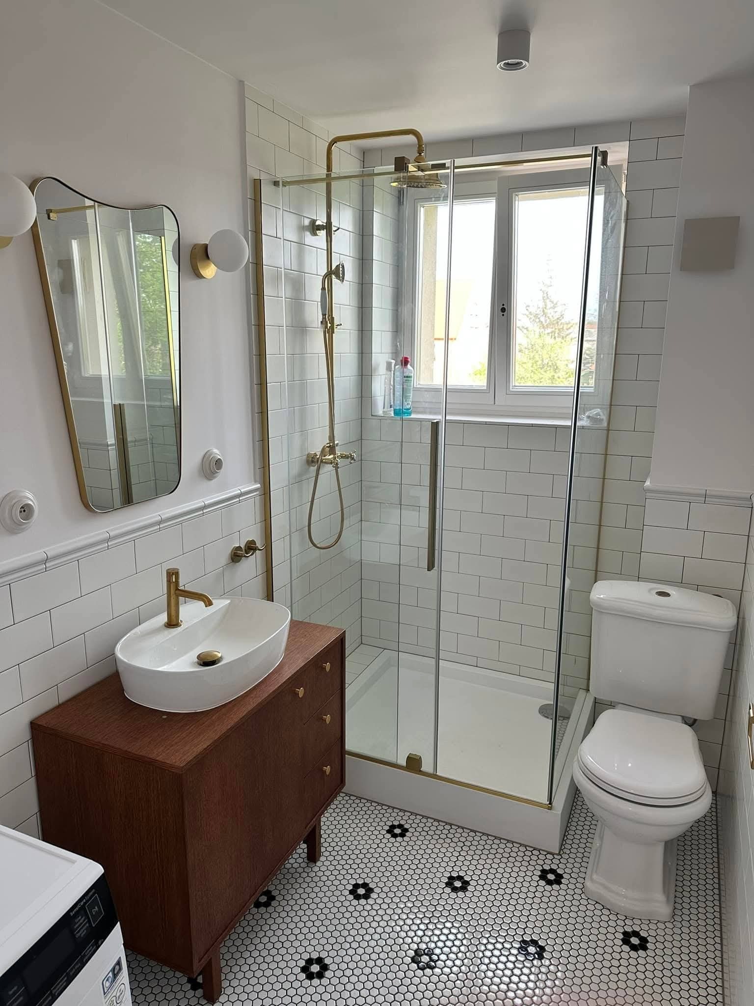Biała łazienka z kabiną prysznicową, umywalką na szafce z drewna i mozaikową podłogą. Armatura w kolorze złotym. Widok na okno.