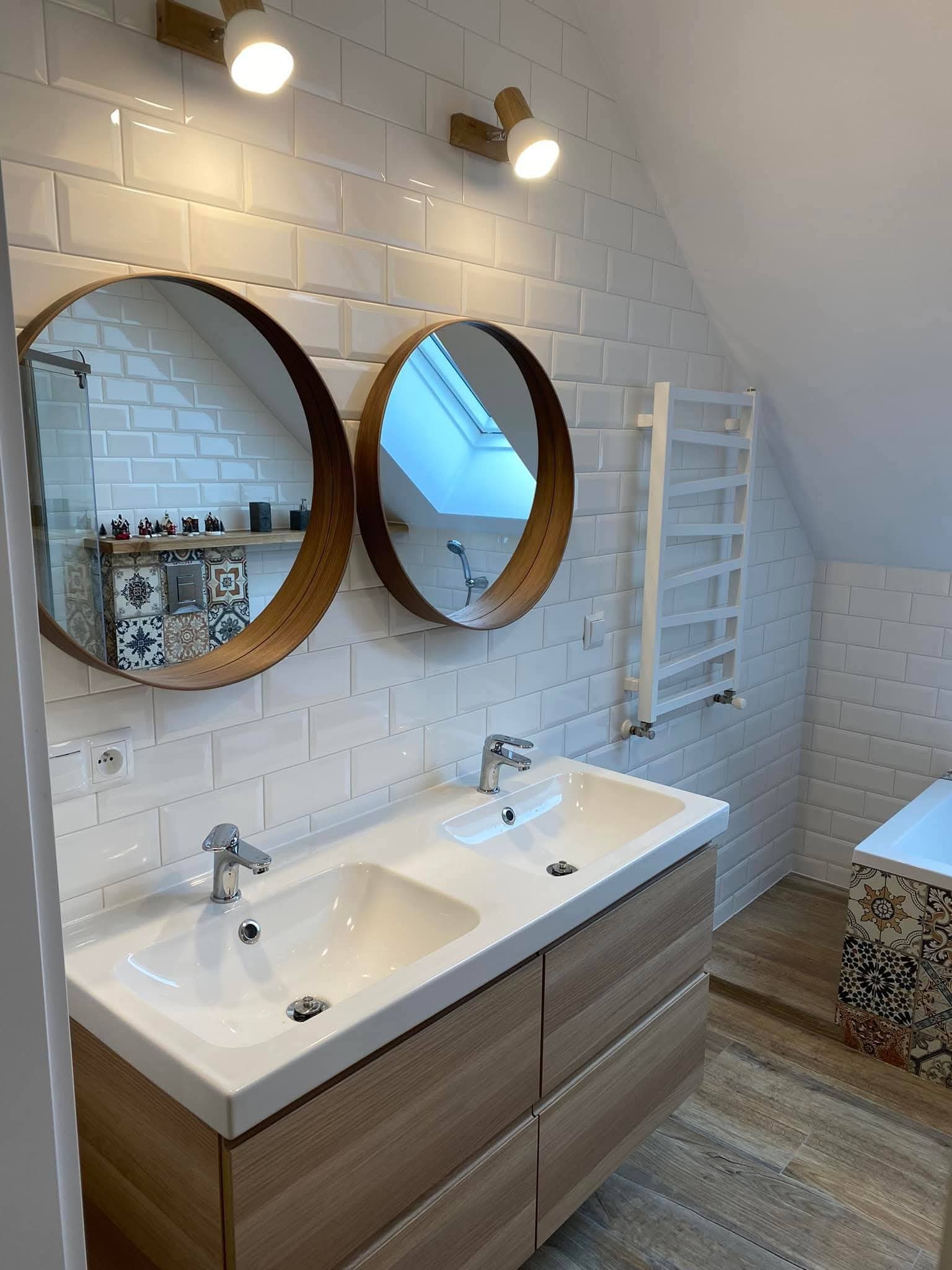 Jasna łazienka na poddaszu z dwoma okrągłymi lustrami, białą armaturą i drewnianymi akcentami. Uwagę przykuwają płytki w stylu metro i dekoracyjne elementy mozaiki.