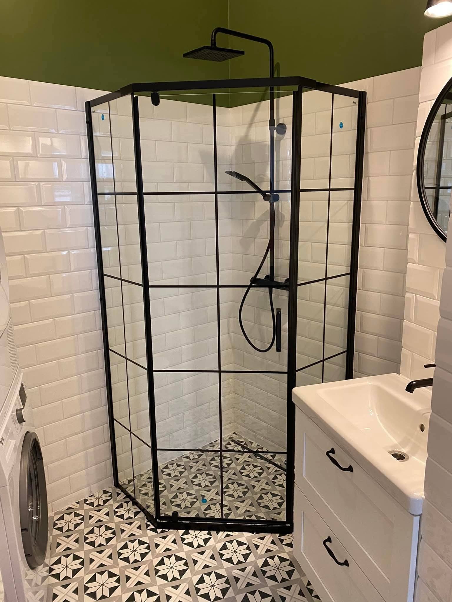 Nowoczesna kabina prysznicowa z czarnymi profilami i geometrycznym wzorem na podłodze w wykończonej łazience z białą umywalką i pralką.