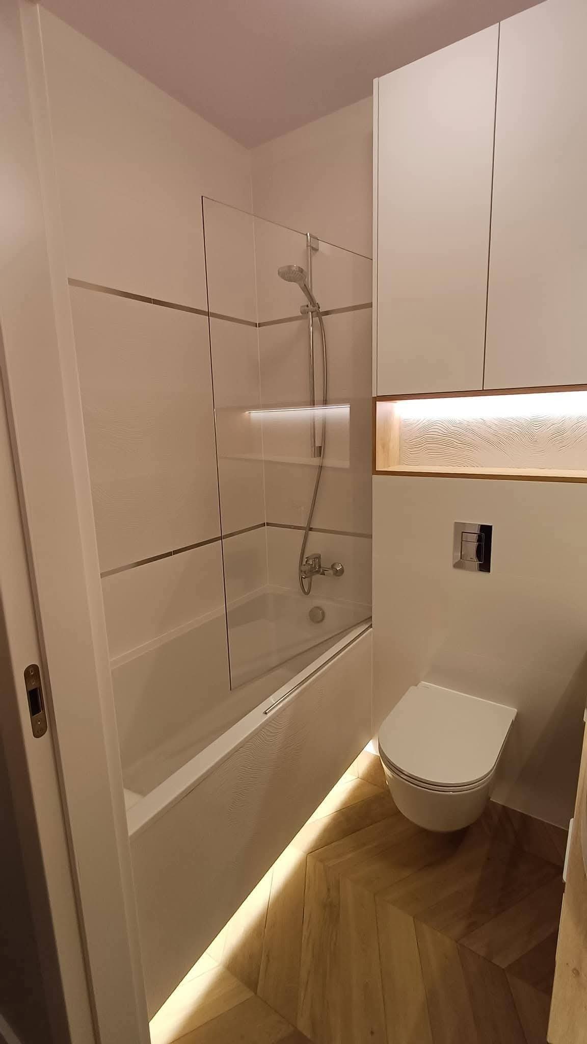 Nowoczesna łazienka z wanną za szklanym parawanem, podświetlaną wnęką i drewnianą podłogą w jodełkę. Minimalistyczny design, jasne kolory, eleganckie wykończenie.