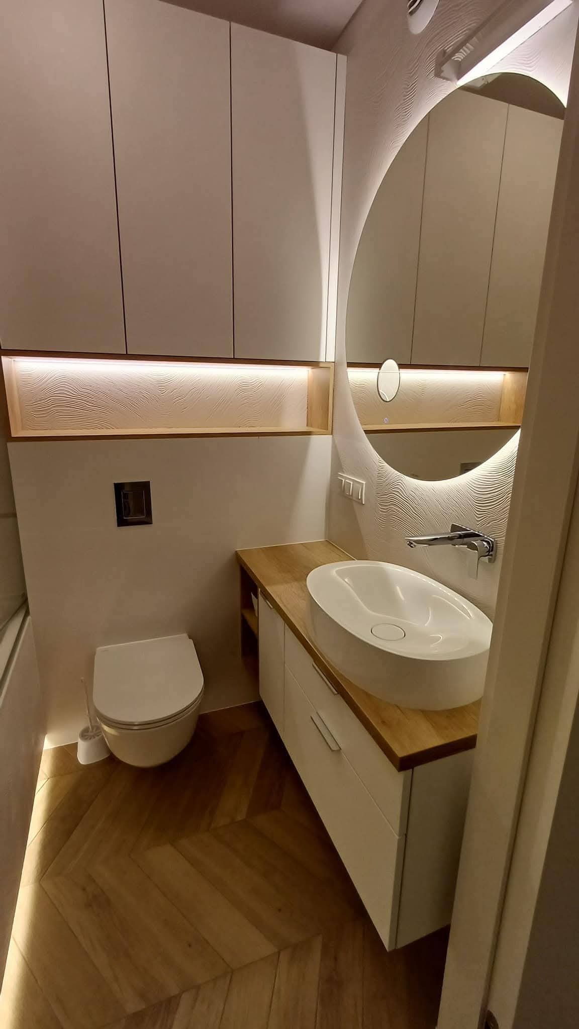 Nowoczesna łazienka z owalną umywalką na drewnianym blacie, podświetlanym lustrem i szafkami w odcieniach bieli. Parkiet ułożony w jodełkę dodaje ciepła.