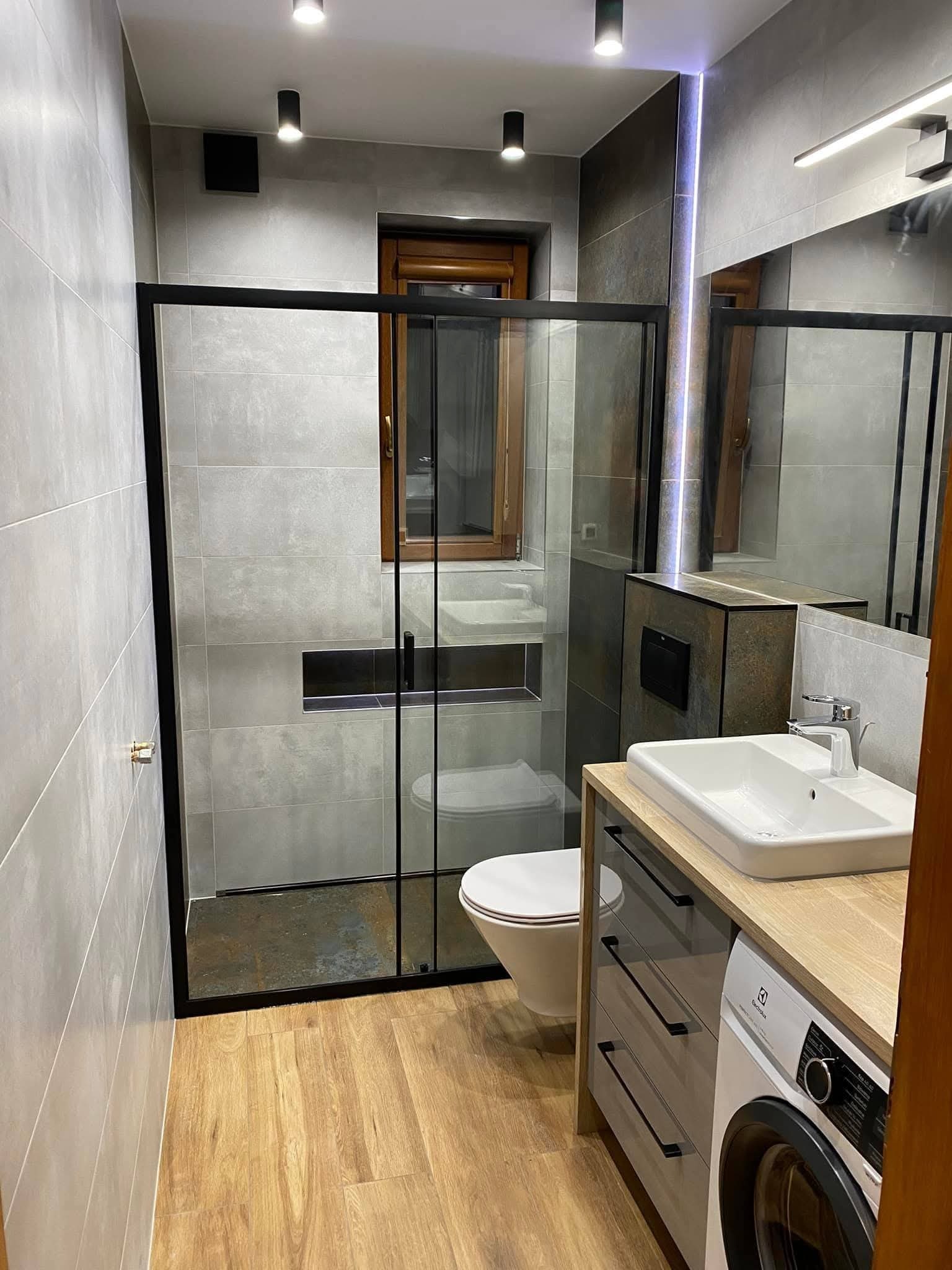 Nowoczesna łazienka z kabiną typu walk-in, szarymi płytkami, drewnopodobną podłogą i pralką zabudowaną pod umywalką. Minimalistyczne oświetlenie punktowe.