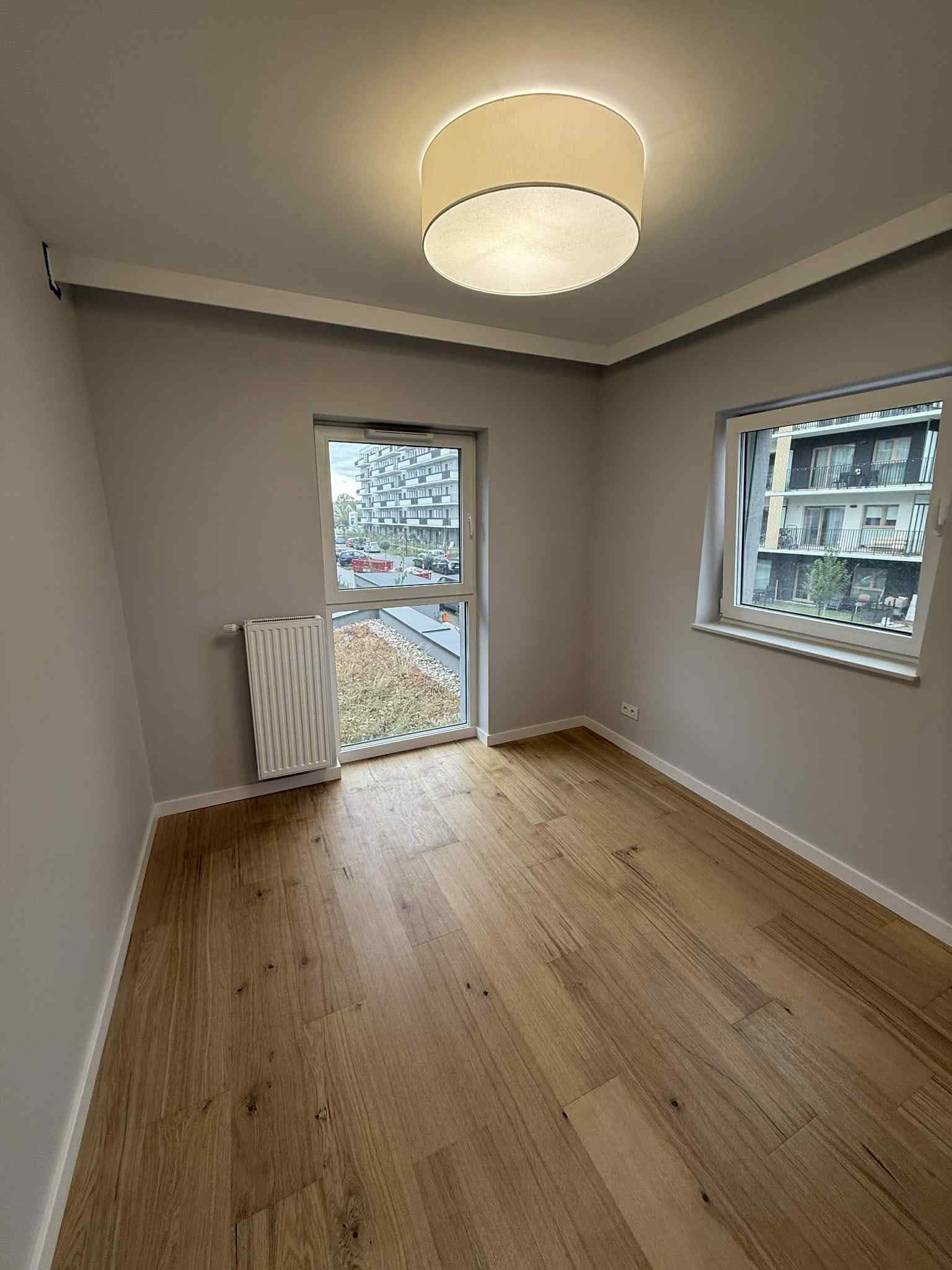 Jasne, drewniane panele podłogowe w pokoju z szarymi ścianami, białymi listwami przypodłogowymi i dwoma oknami z widokiem na zieleń i budynki. Okrągła lampa sufitowa.
