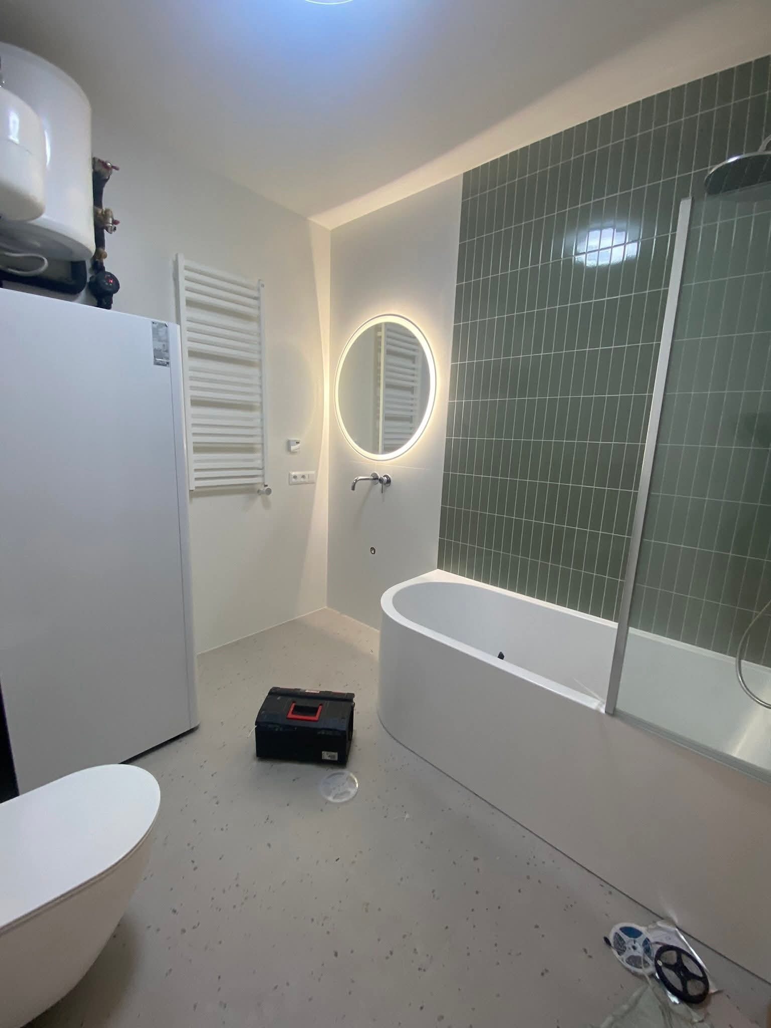 Nowoczesna łazienka po remoncie: zielona mozaika, owalne lustro z podświetleniem, biała wanna z parawanem i grzejnik drabinkowy. Na podłodze leży skrzynka z narzędziami.