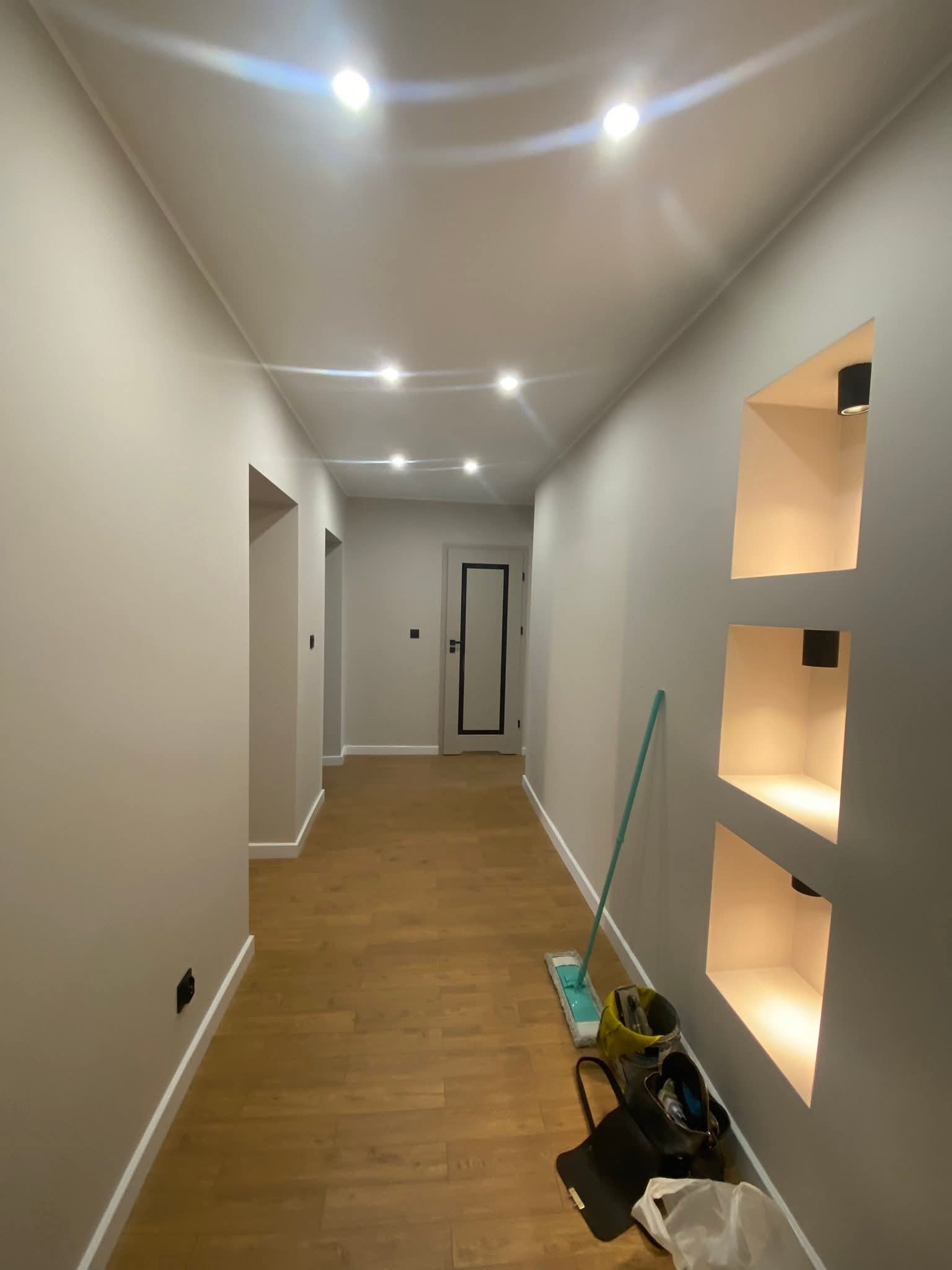 Wykończony korytarz z drewnianą podłogą, wnękami z oświetleniem i świeżo pomalowanymi ścianami w odcieniach szarości. Widoczne akcesoria do sprzątania.