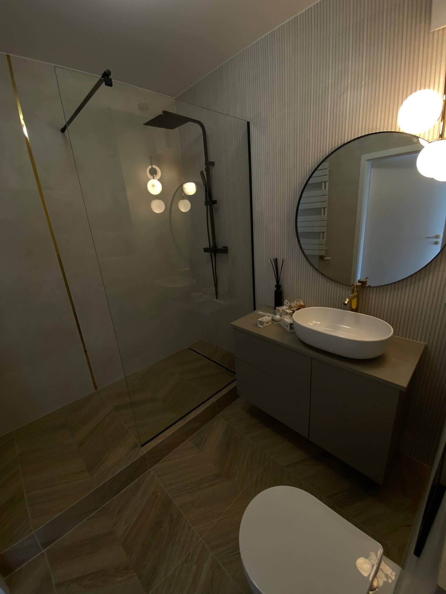 Nowoczesna łazienka z kabiną prysznicową typu walk-in, umywalką nablatową na szafce i okrągłym lustrem. Uwagę zwracają płytki podłogowe ułożone w jodełkę.