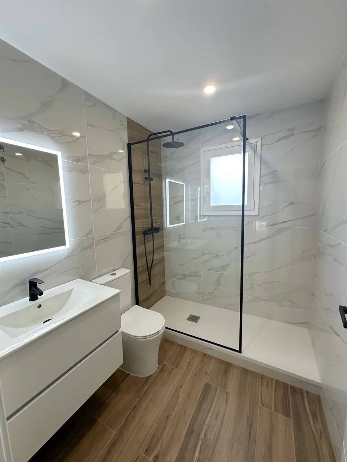 Nowoczesna łazienka po remoncie: białe marmurowe płytki, kabina prysznicowa z czarną ramą, drewnopodobna podłoga, podświetlane lustro i biała szafka z umywalką.