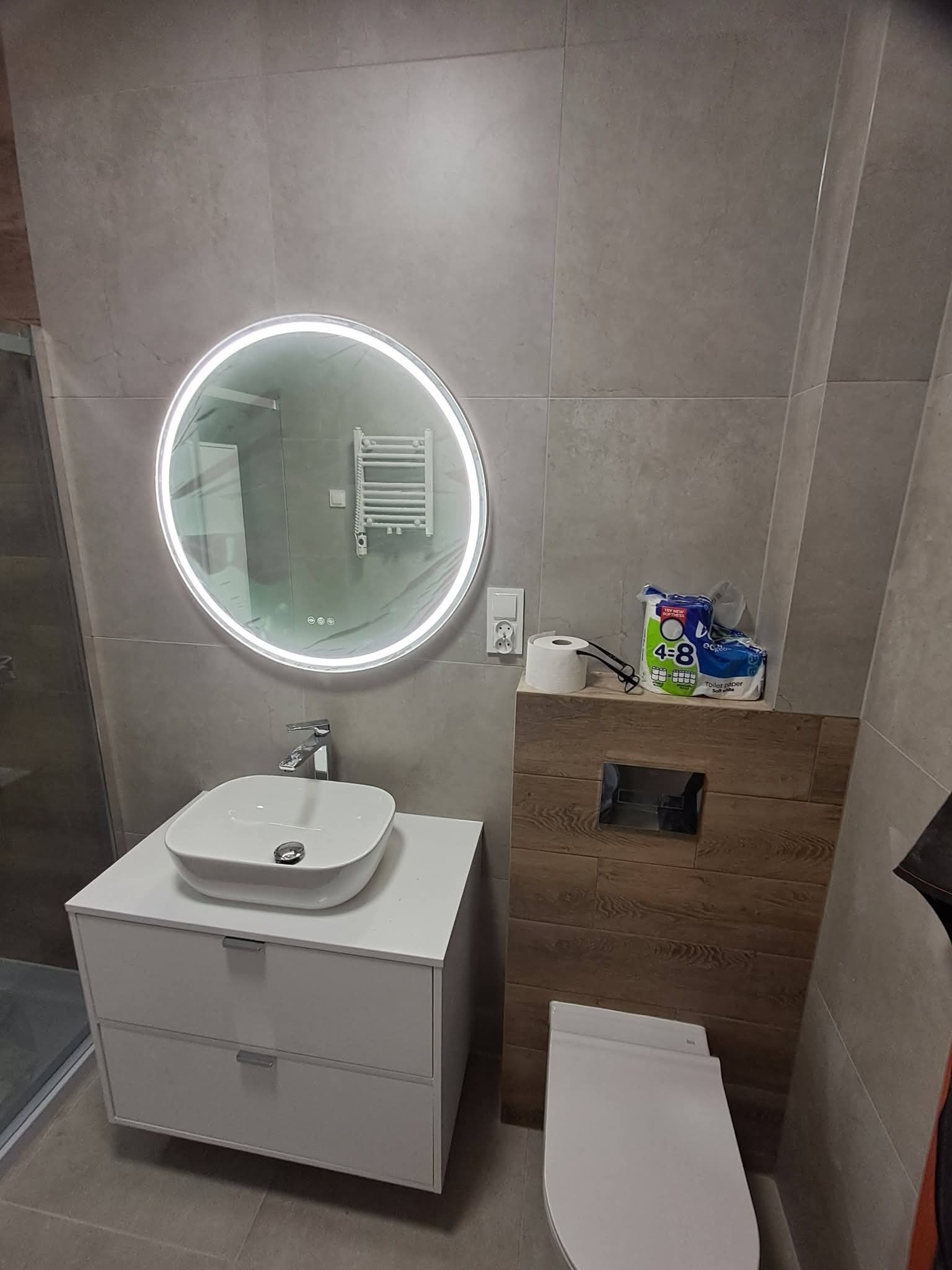 Nowoczesna łazienka z szarą płytką, okrągłym lustrem z podświetleniem LED, białą szafką z umywalką i toaletą. Widoczne akcesoria łazienkowe.
