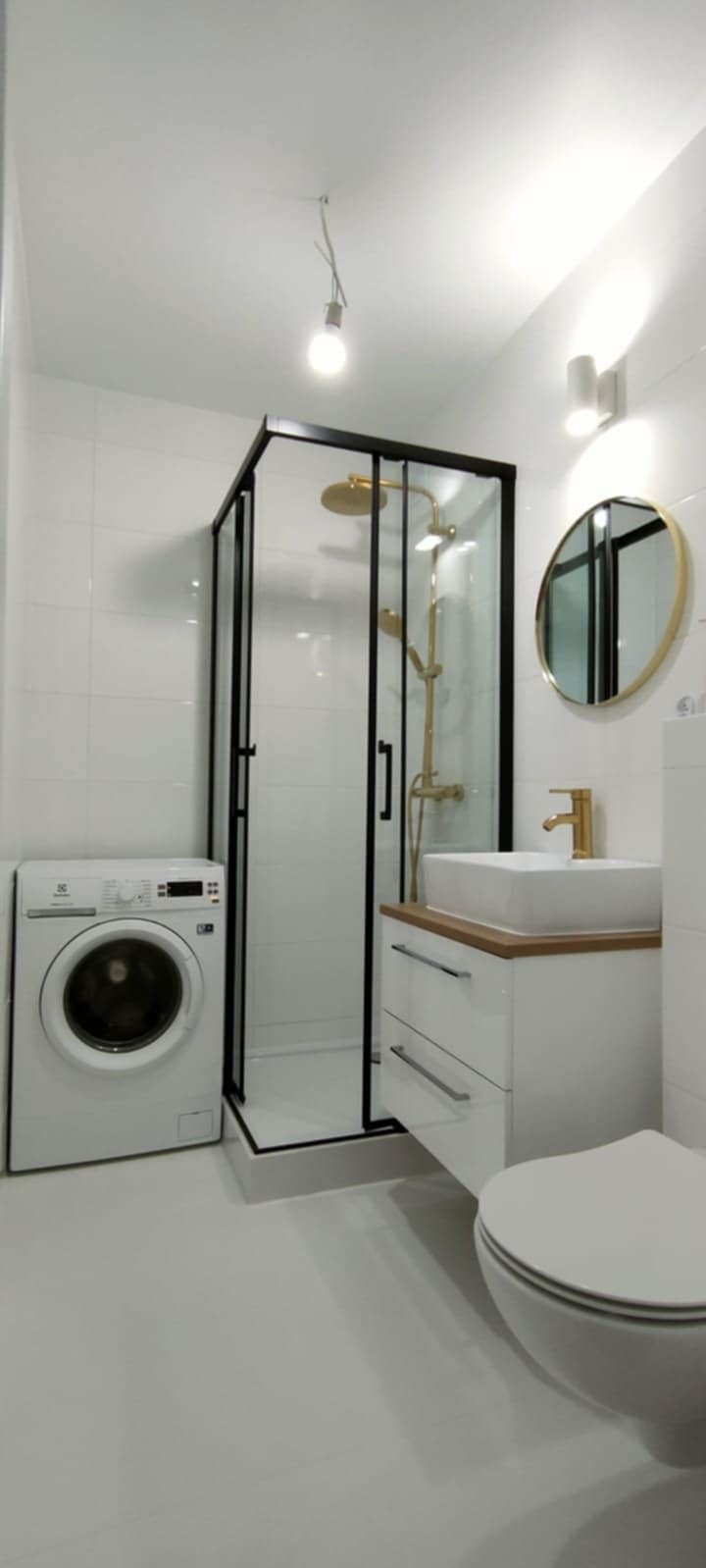 Nowoczesna łazienka w bieli: kabina prysznicowa z czarnymi ramami, złota armatura, okrągłe lustro i pralka. Minimalistyczny design z akcentami złota.
