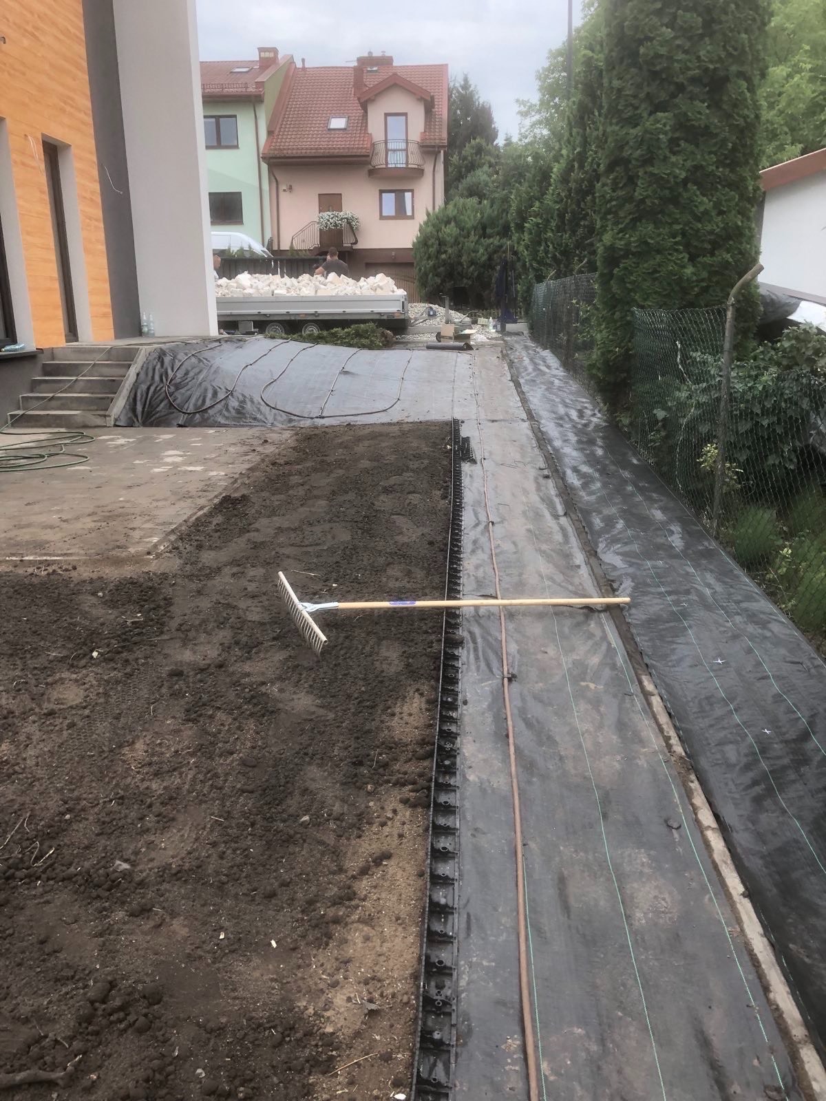 Przygotowany teren pod trawnik z rozłożoną agrowłókniną i systemem nawadniania. Grabie leżą na ziemi, w tle budynek mieszkalny i materiały budowlane.