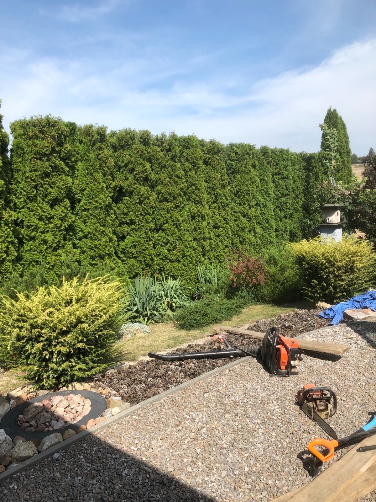 Przygotowany ogród z równo przyciętym żywopłotem, narzędziami ogrodniczymi i wyznaczonym miejscem na ognisko. Widoczny fragment trawnika i niebieska plandeka.