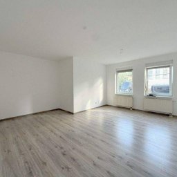 Wardega-budd - Jasne wnętrze po remoncie: panele podłogowe w odcieniach szarości, białe ściany, dwa okna z widokiem na zewnątrz. Minimalistyczny styl, grzejniki pod oknami.