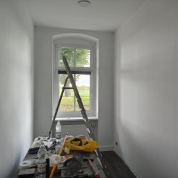 Wardega-budd - Wnętrze po malowaniu na biało, widoczne ściany, okno z widokiem na zieleń, drabina oparta o okno, narzędzia malarskie na stole. Minimalistyczny styl.