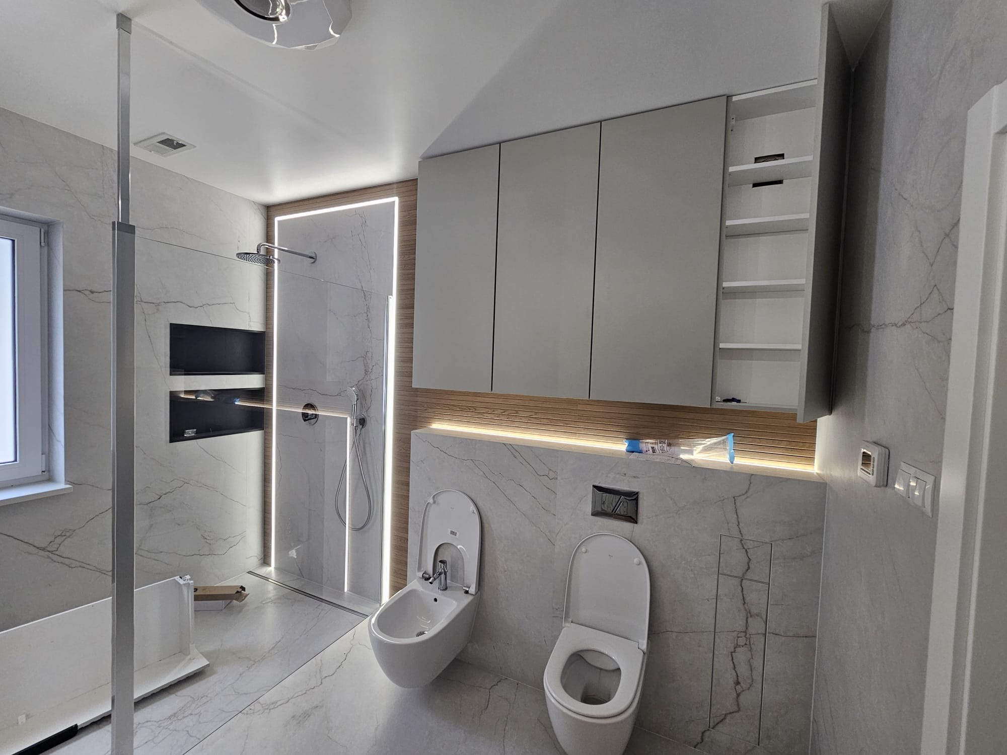 Wykończona łazienka w szarościach z marmurowymi płytkami, podświetlaną kabiną prysznicową typu walk-in, wiszącą szafką i bidetem. Nowoczesny design, czystość i elegancja.
