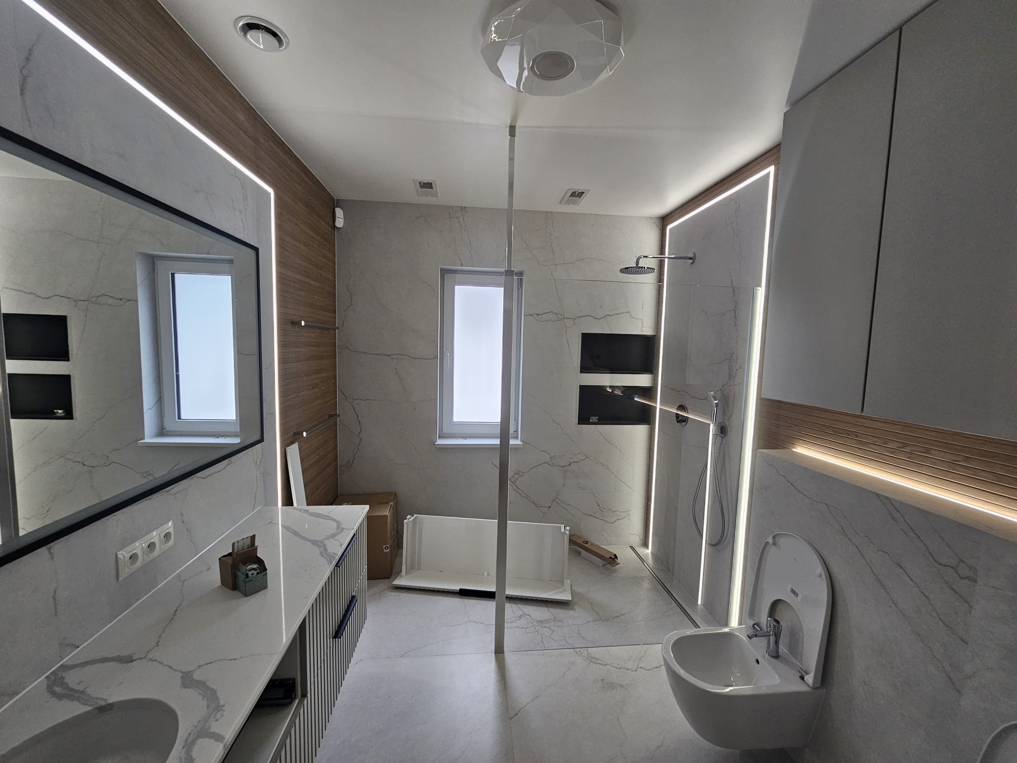 Nowoczesna łazienka w trakcie wykańczania: marmurowe płytki, podświetlane lustro i półki, białe sanitariaty, kabina prysznicowa typu walk-in. Widoczne elementy montażowe.