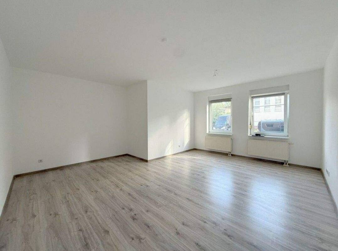 Jasne wnętrze po remoncie: panele podłogowe w odcieniach szarości, białe ściany, dwa okna z widokiem na zewnątrz. Minimalistyczny styl, grzejniki pod oknami.