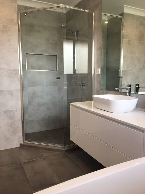 Nowoczesna łazienka z kabiną prysznicową typu walk-in, białą szafką umywalkową i dużym lustrem. Szare płytki na ścianach i ciemna podłoga tworzą elegancki kontrast.