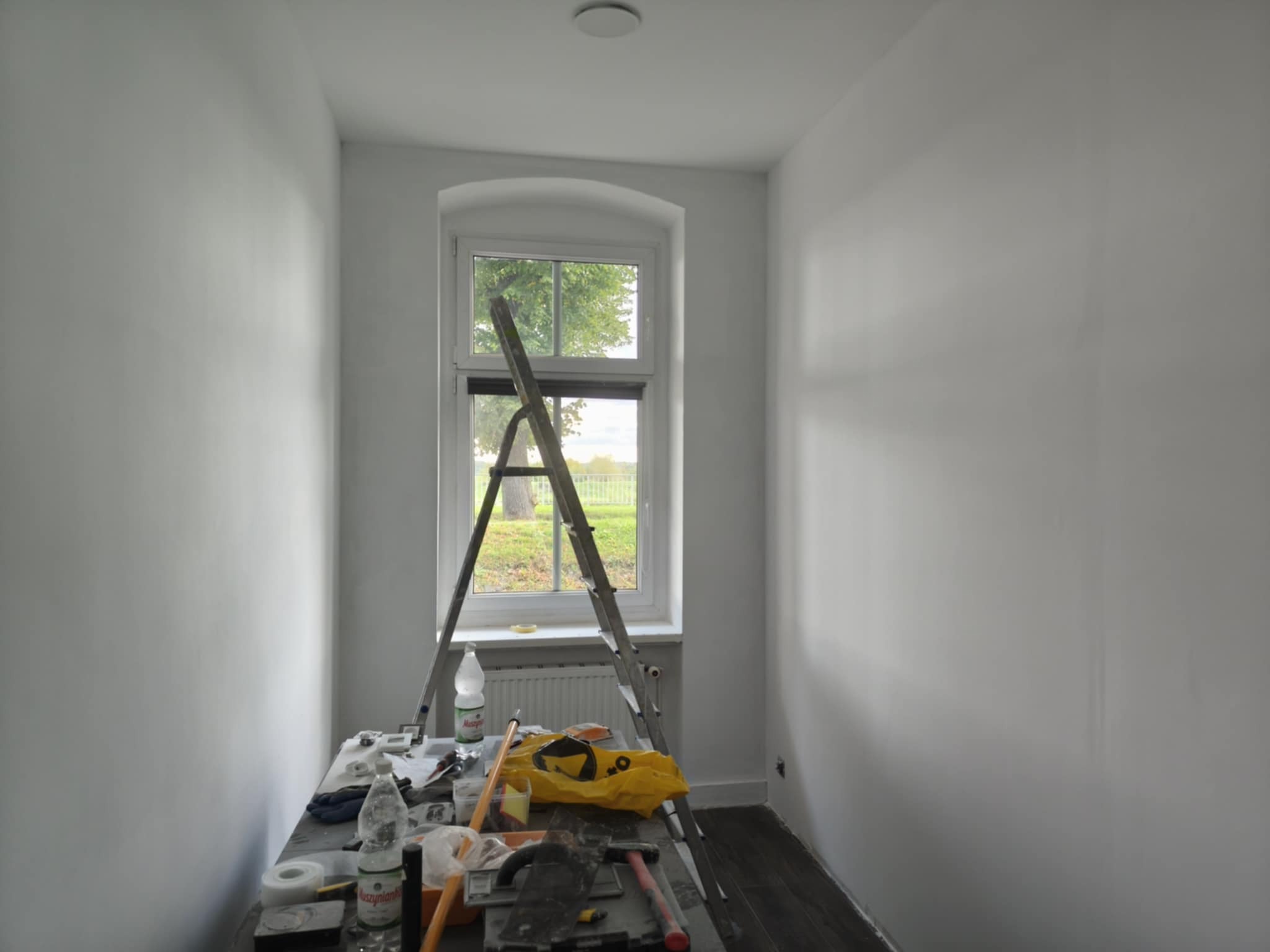 Wnętrze po malowaniu na biało, widoczne ściany, okno z widokiem na zieleń, drabina oparta o okno, narzędzia malarskie na stole. Minimalistyczny styl.