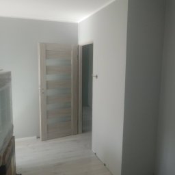Daniel Huczyński - Jasne wnętrze po remoncie: szare ściany, panele podłogowe i nowoczesne drzwi z przeszkleniami. Widoczny fragment mebla zabezpieczonego folią.