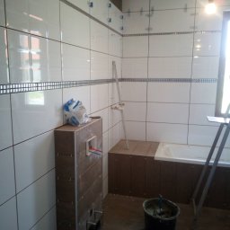 Daniel Huczyński - Łazienka w trakcie remontu: białe płytki z dekoracyjnym paskiem, widoczne krzyżyki dystansowe, zabudowa WC, wiaderko z zaprawą i drabina.