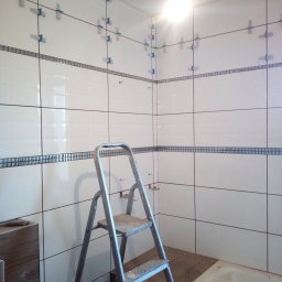 Daniel Huczyński - Białe płytki z dekoracyjnym pasem mozaiki w trakcie układania w łazience, widoczne krzyżyki dystansowe, drabina aluminiowa i fragment wanny.