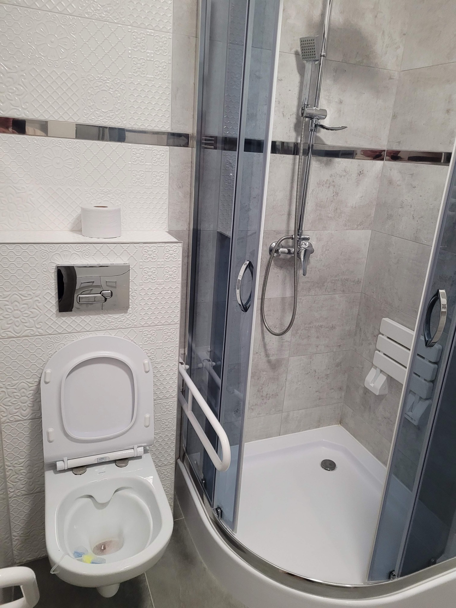 Nowoczesna łazienka z kabiną prysznicową i podwieszaną toaletą. Białe płytki z subtelnym wzorem. Chromowane detale. Umywalka z baterią. Minimalistyczny design.