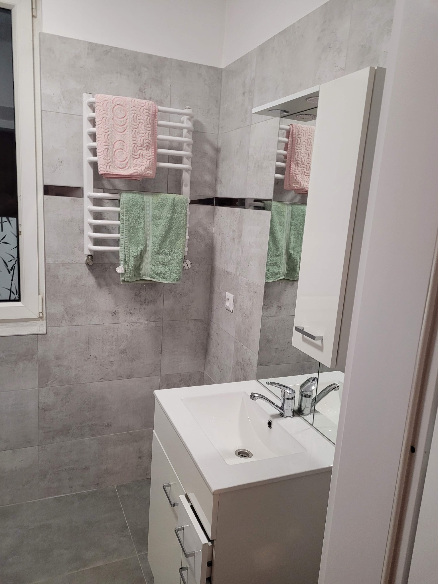 Jasna łazienka z szarymi płytkami, białą szafką z umywalką i grzejnikiem drabinkowym z ręcznikami. Widoczne okno i lustro z szafką.