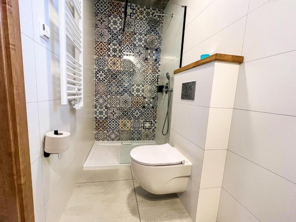 Nowoczesna łazienka z kabiną prysznicową wykończoną mozaiką, podwieszaną toaletą i grzejnikiem drabinkowym. Białe płytki i drewniane akcenty.