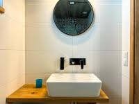 Nowoczesna łazienka: biała, prostokątna umywalka na drewnianym blacie, okrągłe lustro i minimalistyczna armatura na tle płytek w odcieniach beżu.