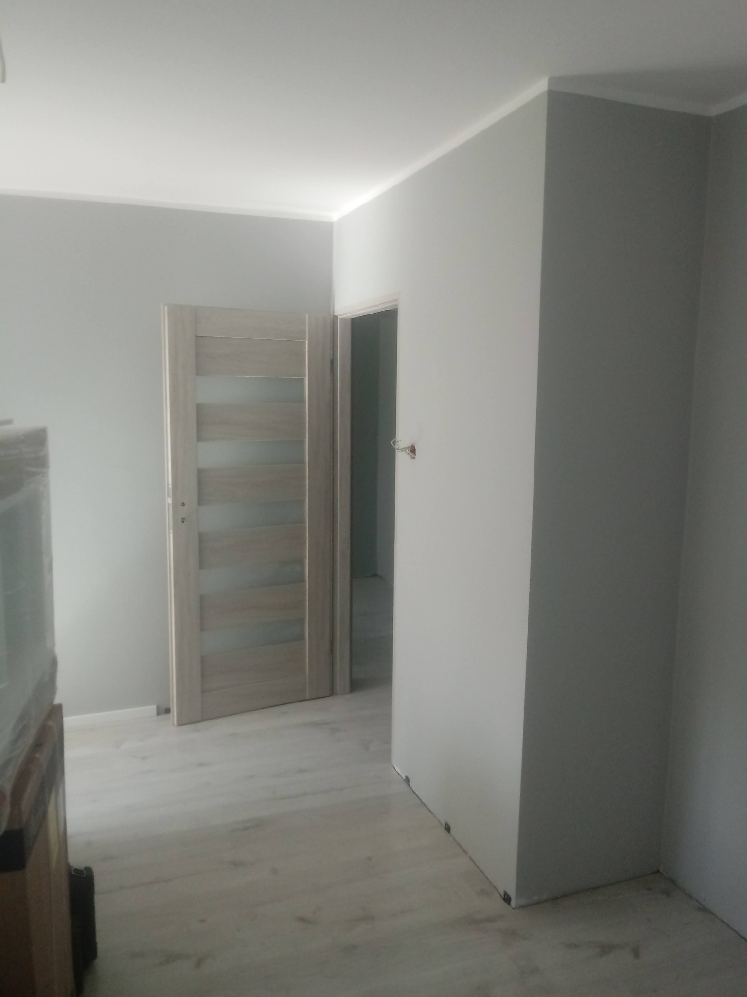 Jasne wnętrze po remoncie: szare ściany, panele podłogowe i nowoczesne drzwi z przeszkleniami. Widoczny fragment mebla zabezpieczonego folią.