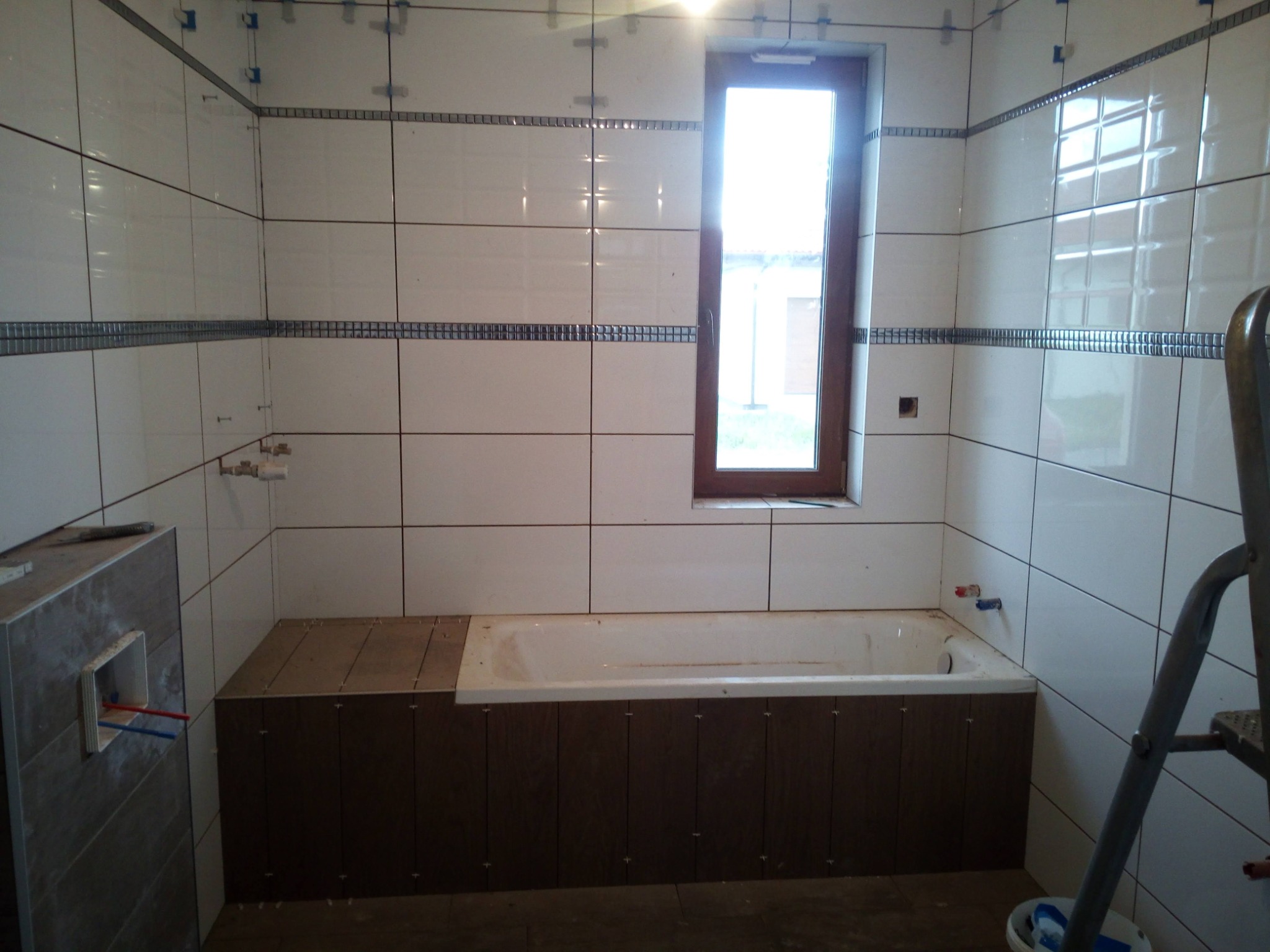 Łazienka w trakcie wykańczania: białe płytki z dekoracyjnym pasem, obudowana wanna, widoczne krzyżyki glazurnicze, drabina. Okno z widokiem na zewnątrz.