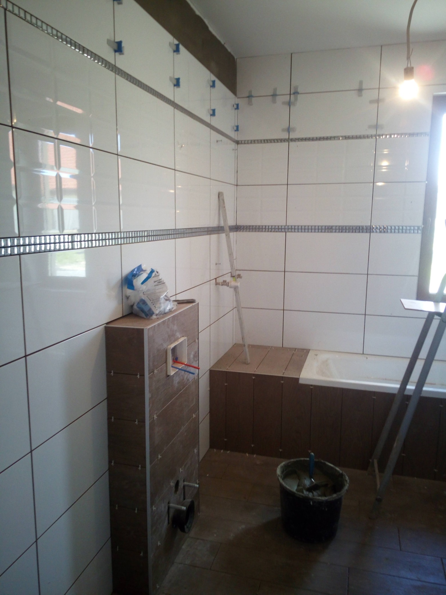 Łazienka w trakcie remontu: białe płytki z dekoracyjnym paskiem, widoczne krzyżyki dystansowe, zabudowa WC, wiaderko z zaprawą i drabina.