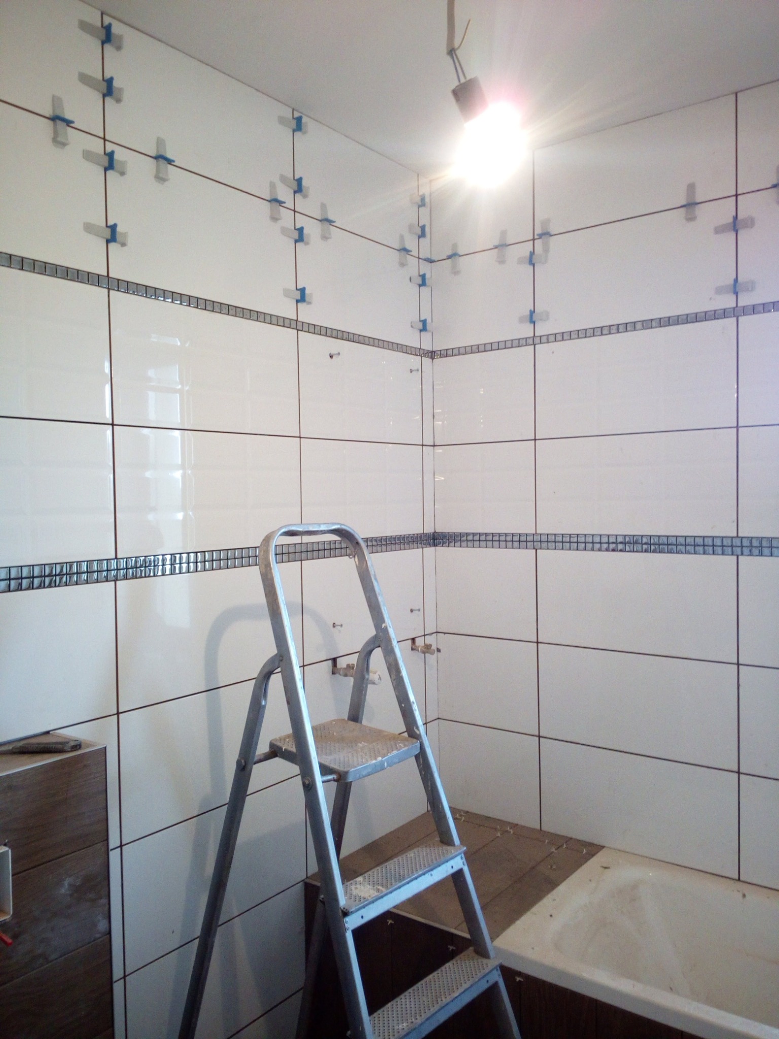 Białe płytki z dekoracyjnym pasem mozaiki w trakcie układania w łazience, widoczne krzyżyki dystansowe, drabina aluminiowa i fragment wanny.