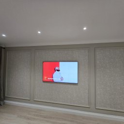 D PAUL LTD - Salon z telewizorem na ścianie w dekoracyjnych ramach. Ściany w odcieniach szarości z subtelnym wzorem. Parkiet na podłodze i zasłony po lewej stronie. Oświetlenie sufitowe punktowe.