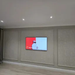 Salon z telewizorem na ścianie w dekoracyjnych ramach. Ściany w odcieniach szarości z subtelnym wzorem. Parkiet na podłodze i zasłony po lewej stronie. Oświetlenie sufitowe punktowe.