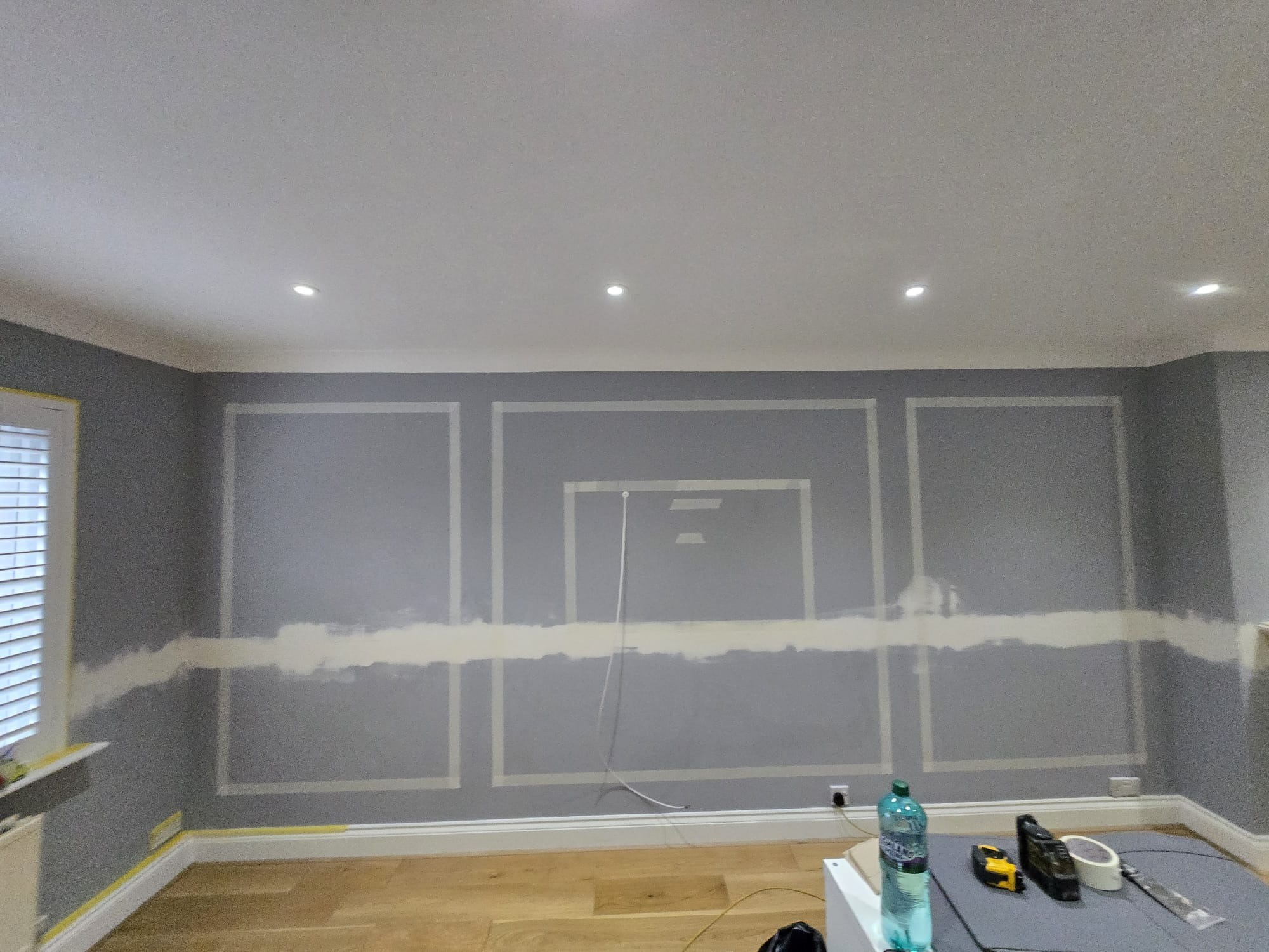 Szara ściana z dekoracyjnymi listwami, częściowo zamaskowana taśmą malarską i wstępnie szpachlowana, przygotowana do dalszych prac wykończeniowych.