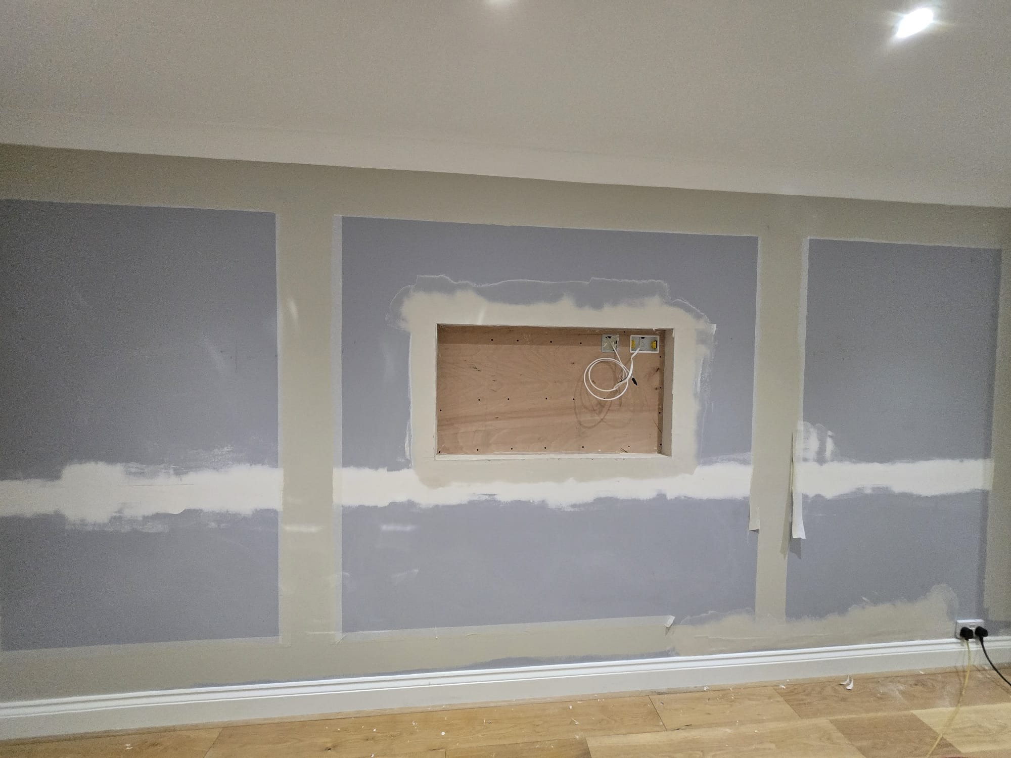 Ściana w trakcie przygotowania do malowania z wnęką na telewizor. Widoczne szpachlowanie i wstępne malowanie na szaro. Podłoga drewniana, listwy przypodłogowe.