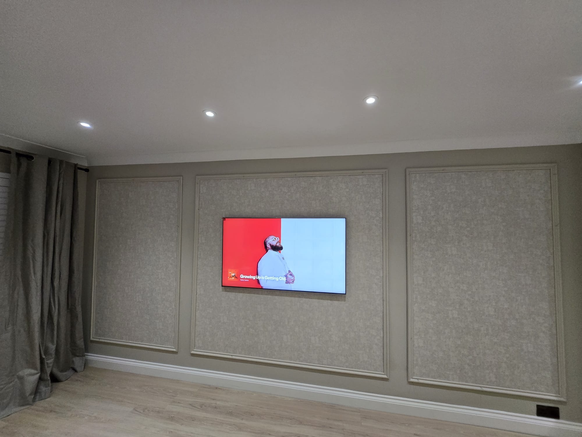 Salon z telewizorem na ścianie w dekoracyjnych ramach. Ściany w odcieniach szarości z subtelnym wzorem. Parkiet na podłodze i zasłony po lewej stronie. Oświetlenie sufitowe punktowe.