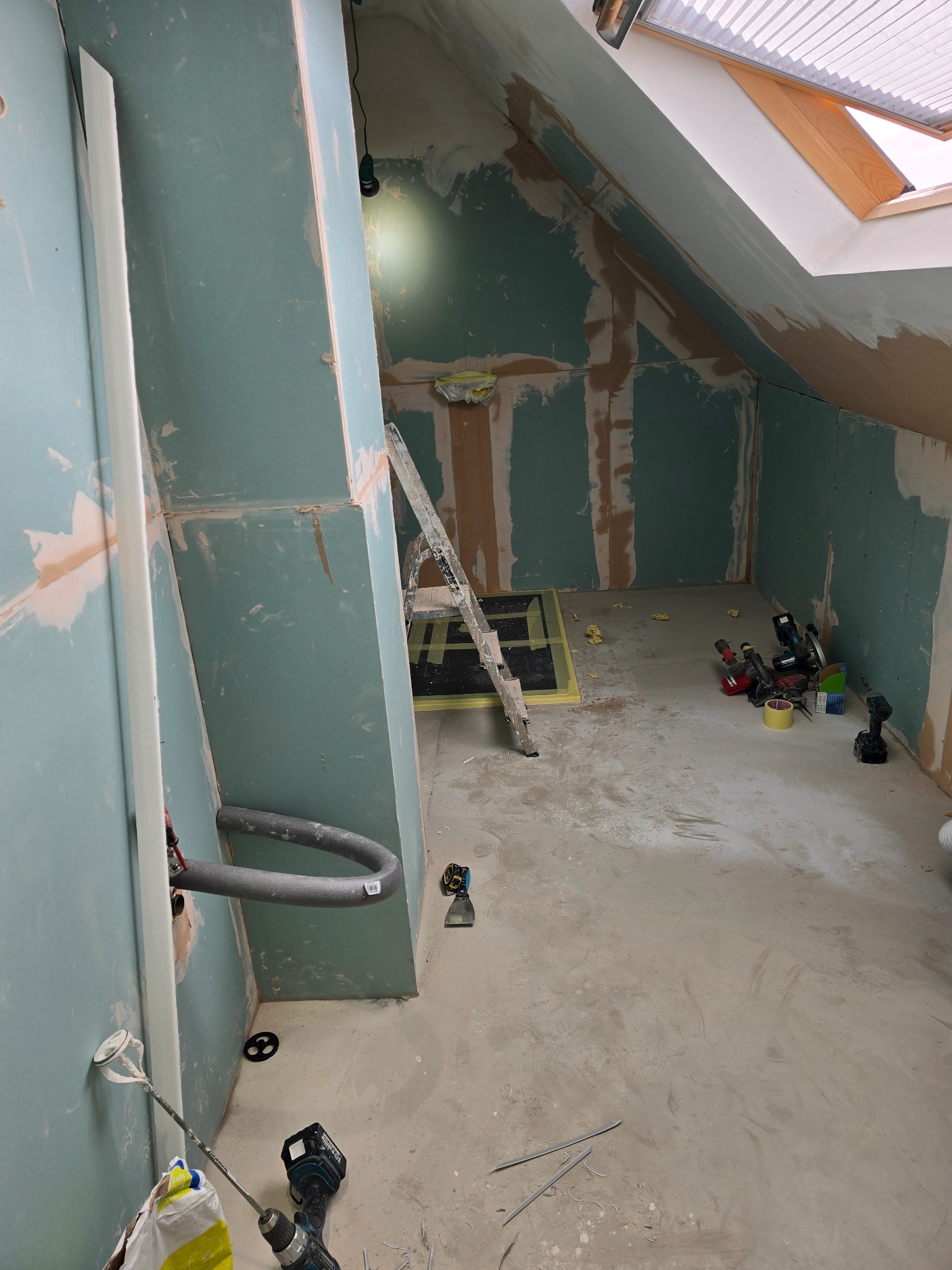 Poddasze w trakcie prac wykończeniowych: płyty gipsowe na ścianach, częściowo zaszpachlowane, narzędzia pracy, drabina i okno dachowe. Widok na surowy stan wnętrza.