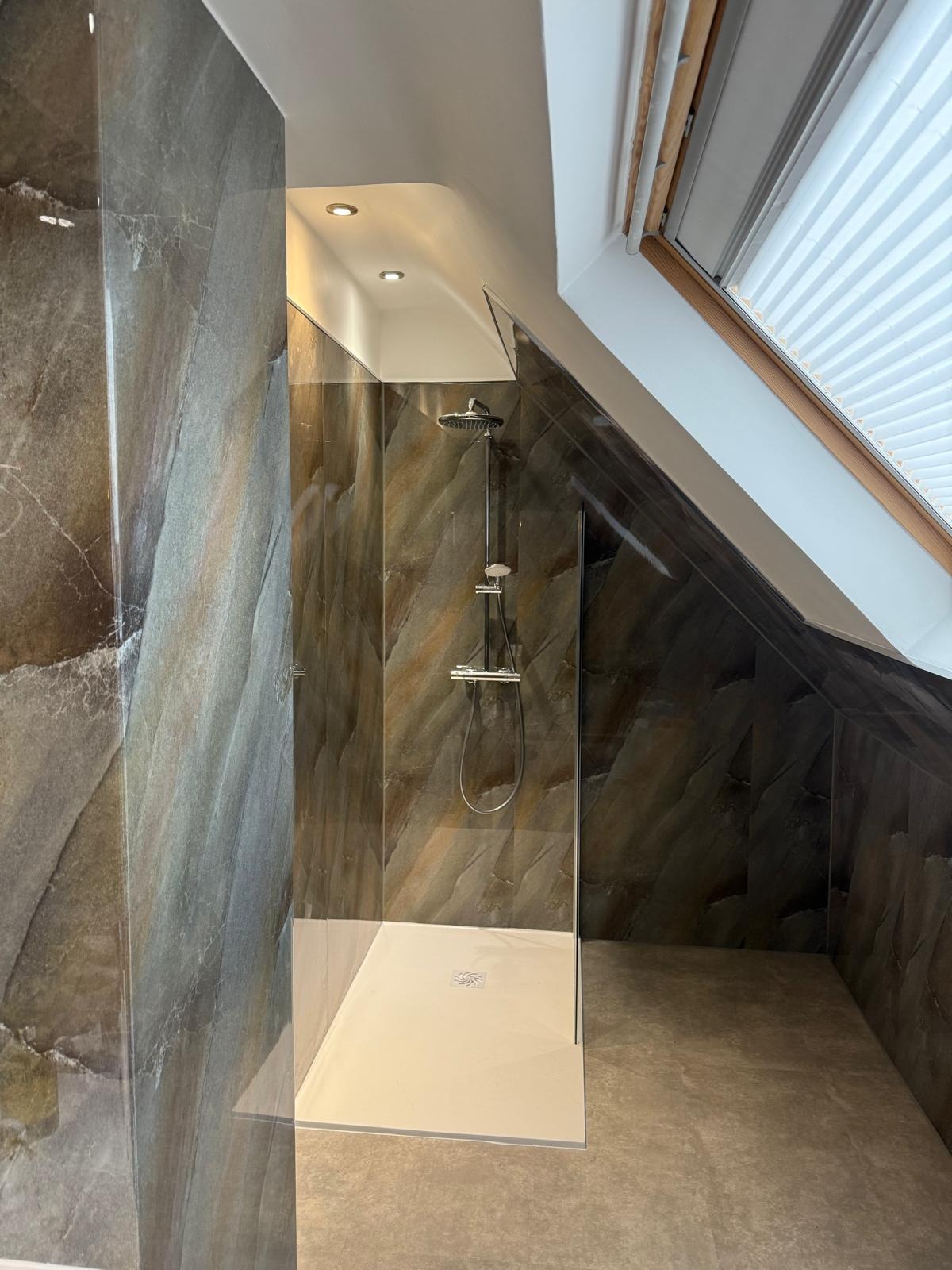 Nowoczesna kabina prysznicowa z deszczownicą w łazience na poddaszu, wykończona płytkami imitującymi marmur. Widoczne okno dachowe z żaluzjami.