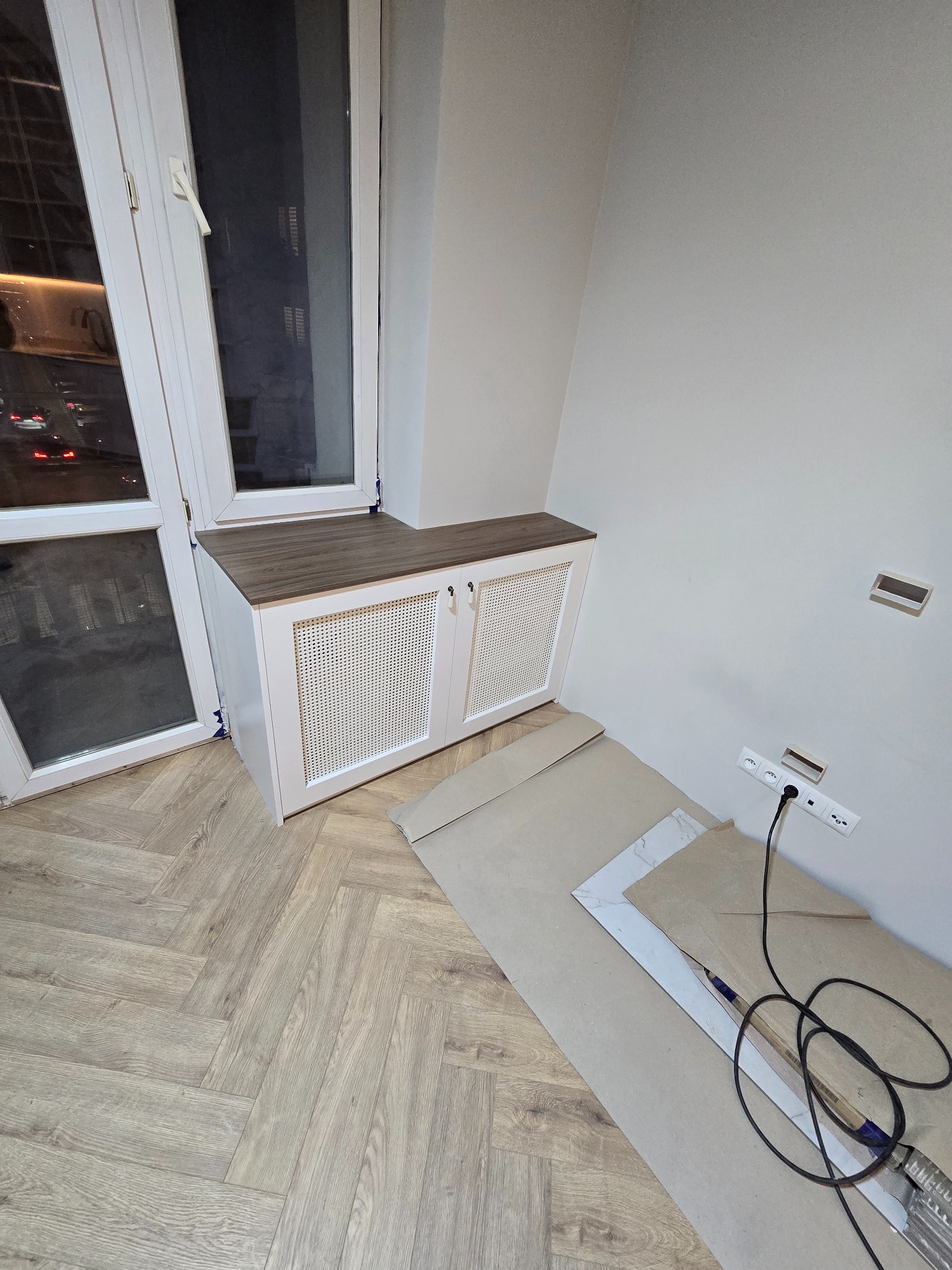 Biała zabudowa grzejnika z drewnianym blatem, dopasowana do okna, na podłodze panele ułożone w jodełkę. Widoczne gniazdka elektryczne i materiały budowlane.