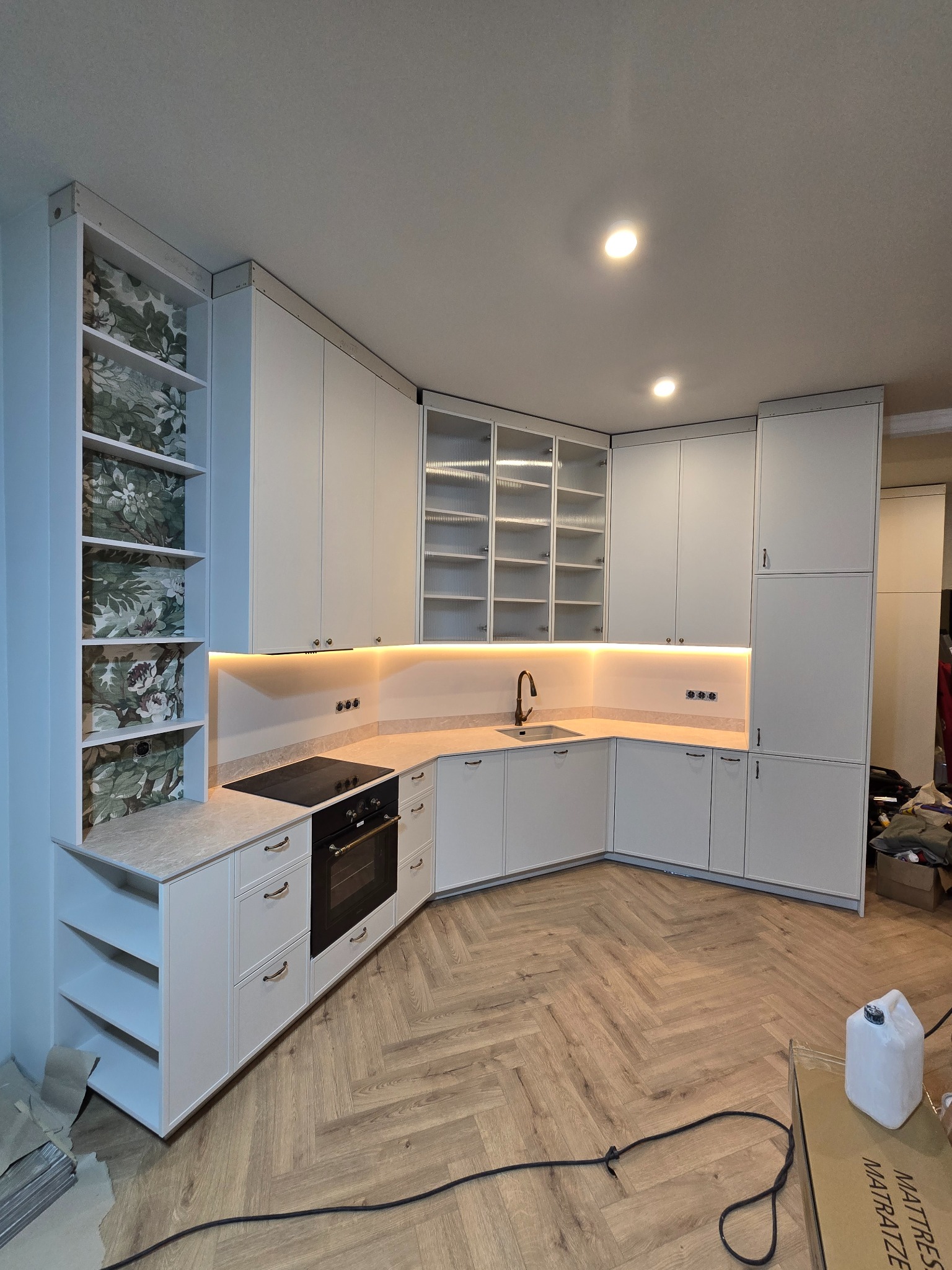 Jasna kuchnia w stylu klasycznym z drewnianą podłogą w jodełkę. Szafki z przeszklonymi witrynami i oświetleniem LED. Na półkach regału wzorzysta tapeta.