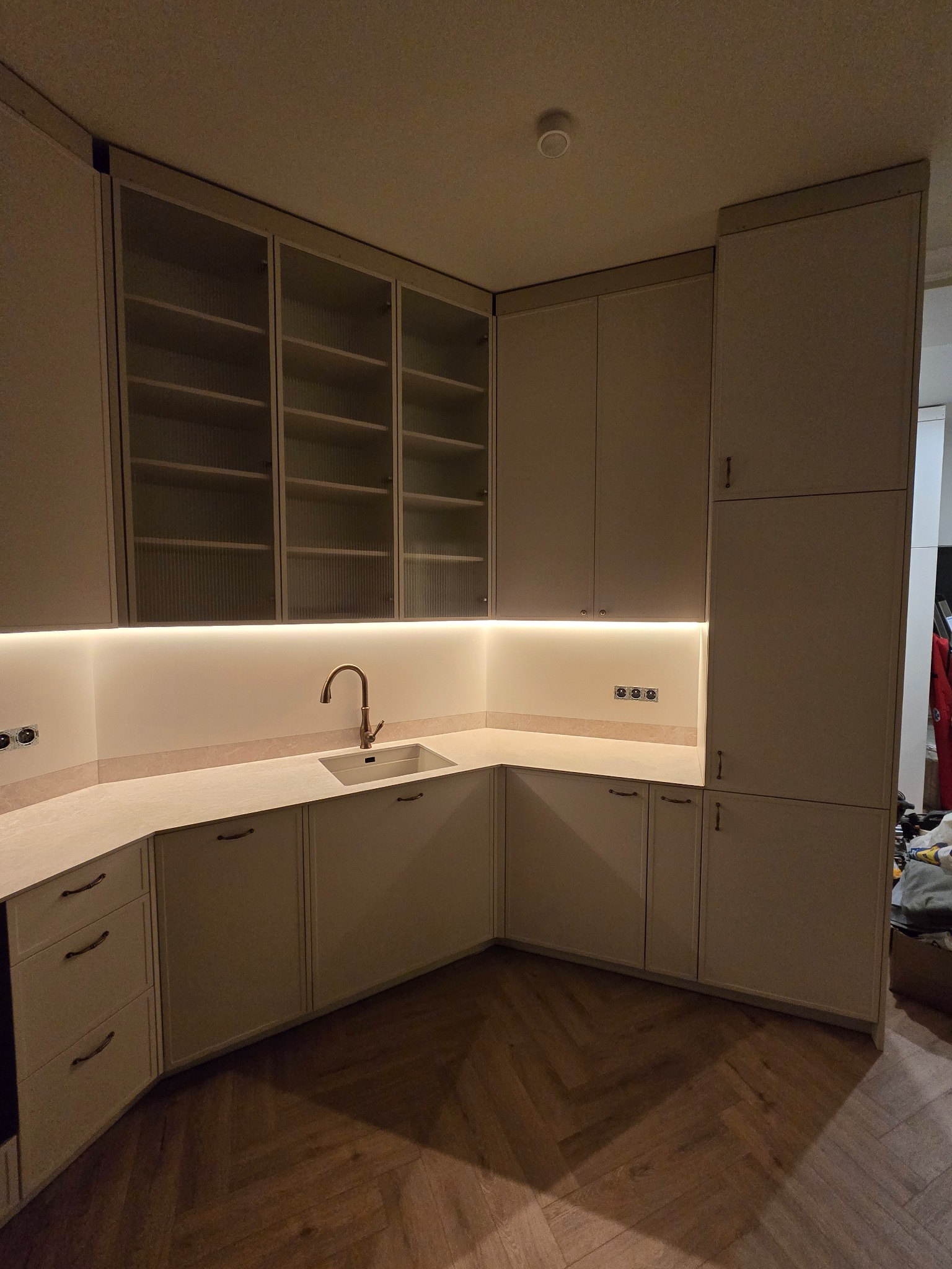 Nowoczesna kuchnia w odcieniach beżu z drewnianą podłogą w jodełkę. Szafki z ryflowanymi szybami, oświetlenie LED pod szafkami wiszącymi, złota bateria.