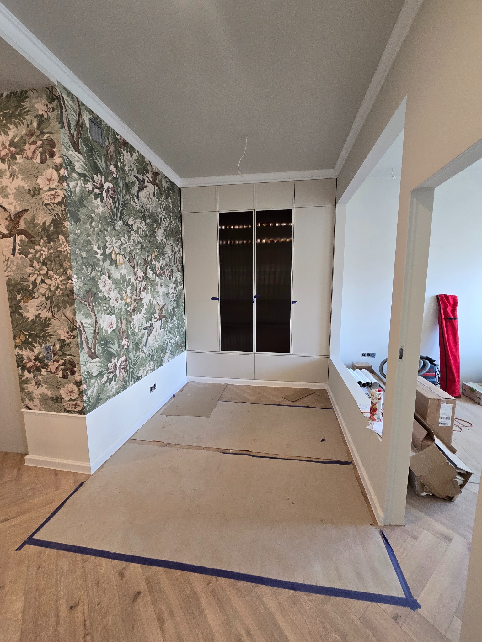 Pokój w trakcie wykańczania: wzorzysta tapeta z motywem roślinnym, zabudowana szafa z ciemnymi szybami, zabezpieczona podłoga. Widoczne kartony i narzędzia.